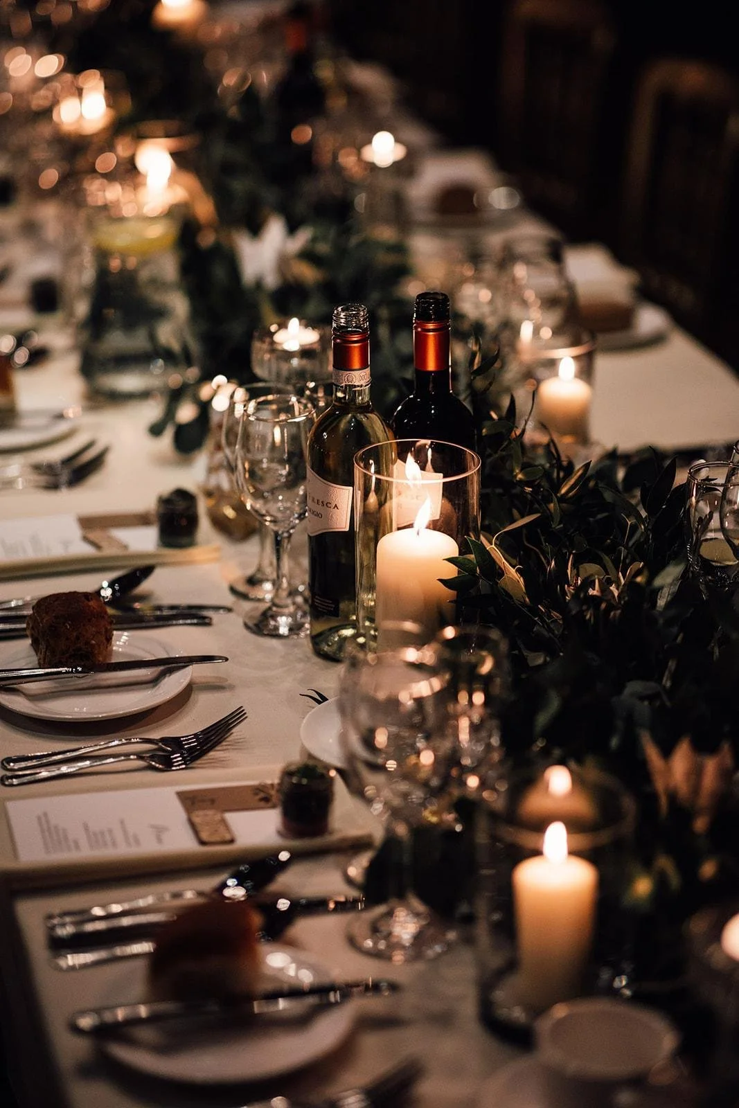 A beautifully set dinner table decorated with candles, wine bottles, glasses, and leafy greenery in a warm, dimly lit ambiance.