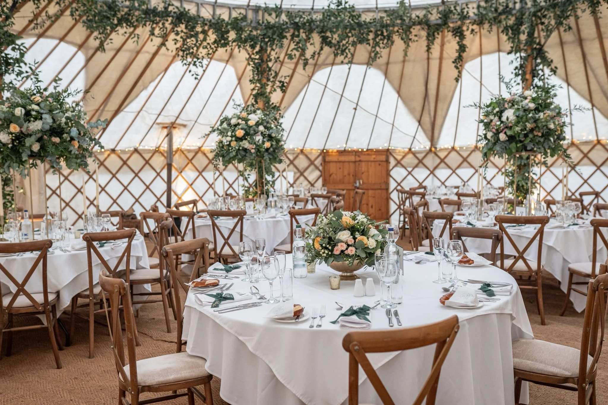 Elegant wedding reception setup inside a large wooden tent, decorated with flowers, greenery, and string lights. Multiple round tables are covered with white tablecloths, adorned with floral centerpieces, glassware, and cutlery.