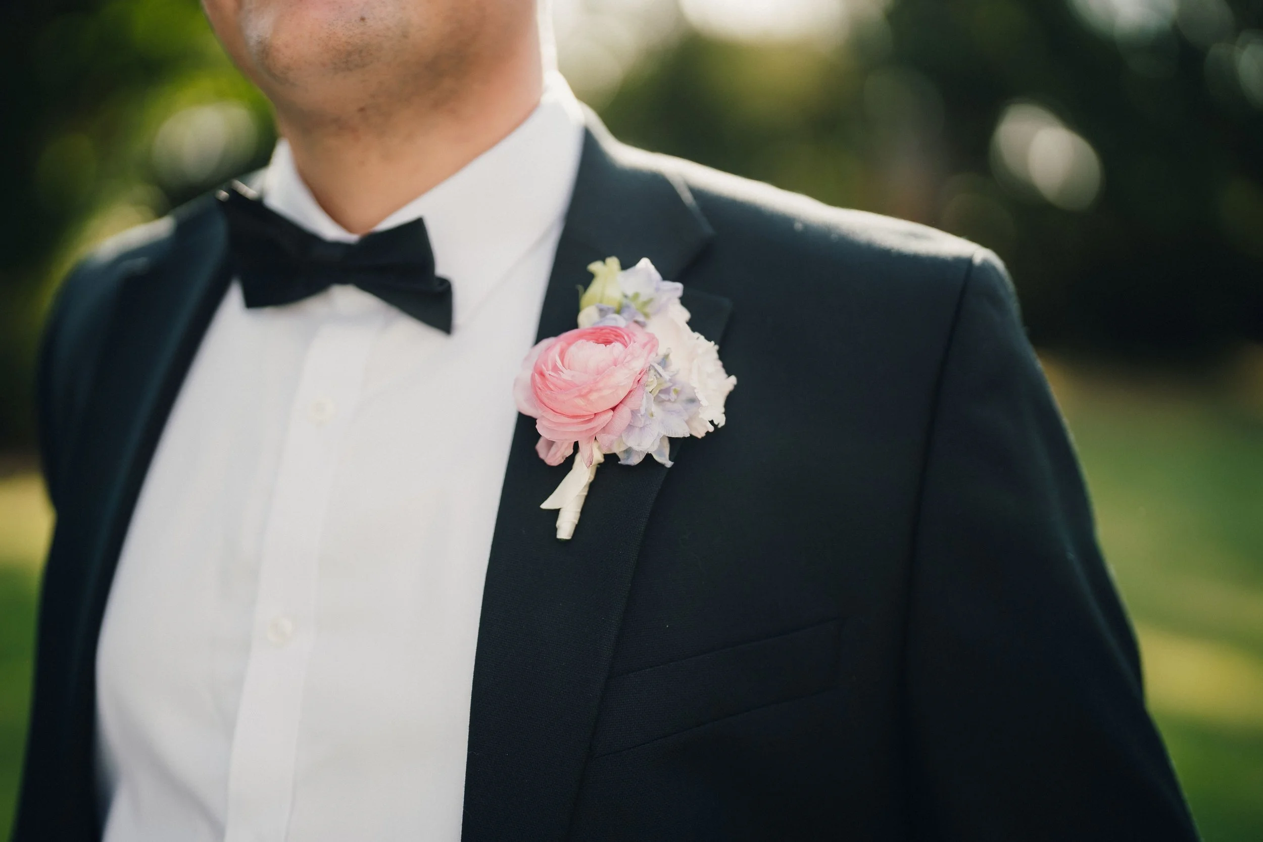 Close-up of a man in formal attire wearing a black tuxedo, white shirt, black bow tie, and a pink and purple boutonniere on his lapel, outdoors with blurred greenery in the background.