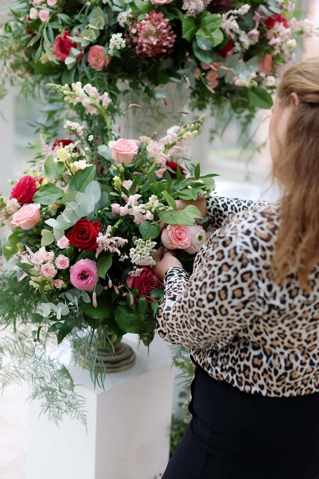 A woman arranging a large pink, red, and white floral bouquet in a white vase, wearing a leopard print blouse.