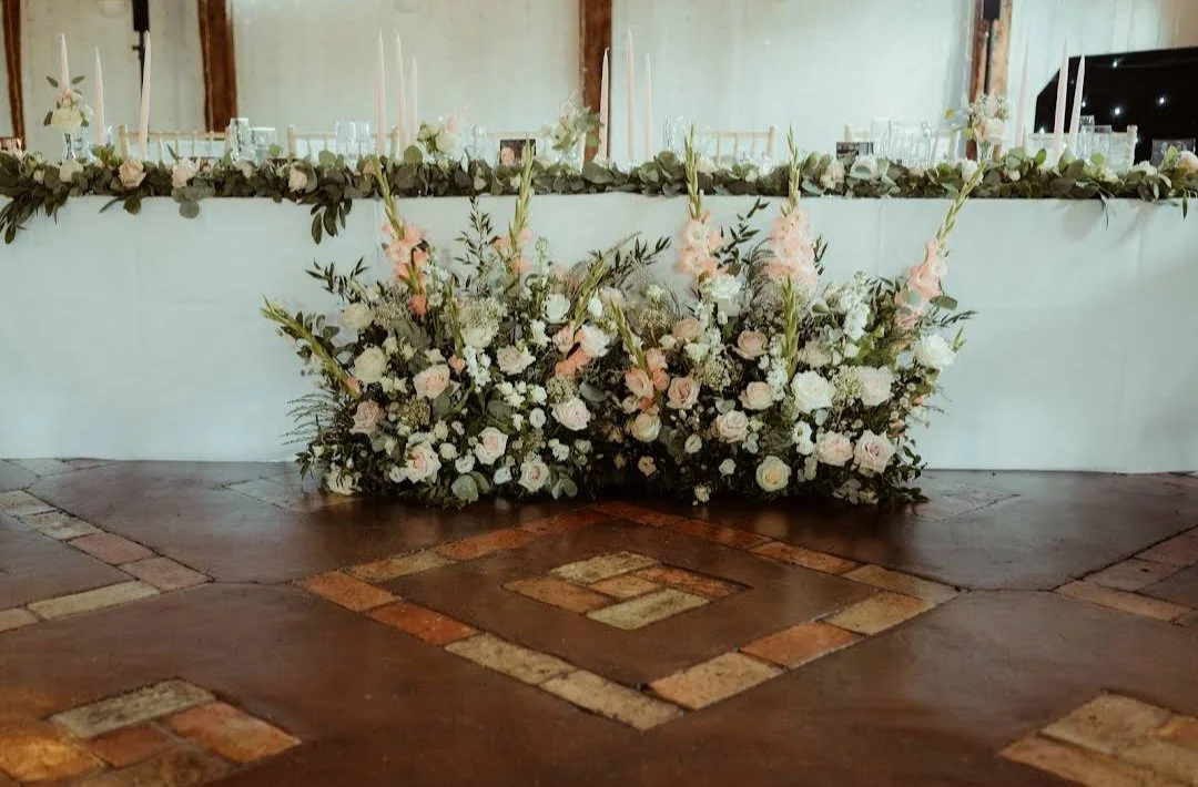 Flower arrangement with white roses and pink gladiolus on a brick floor in front of a long table decorated with a floral garland.