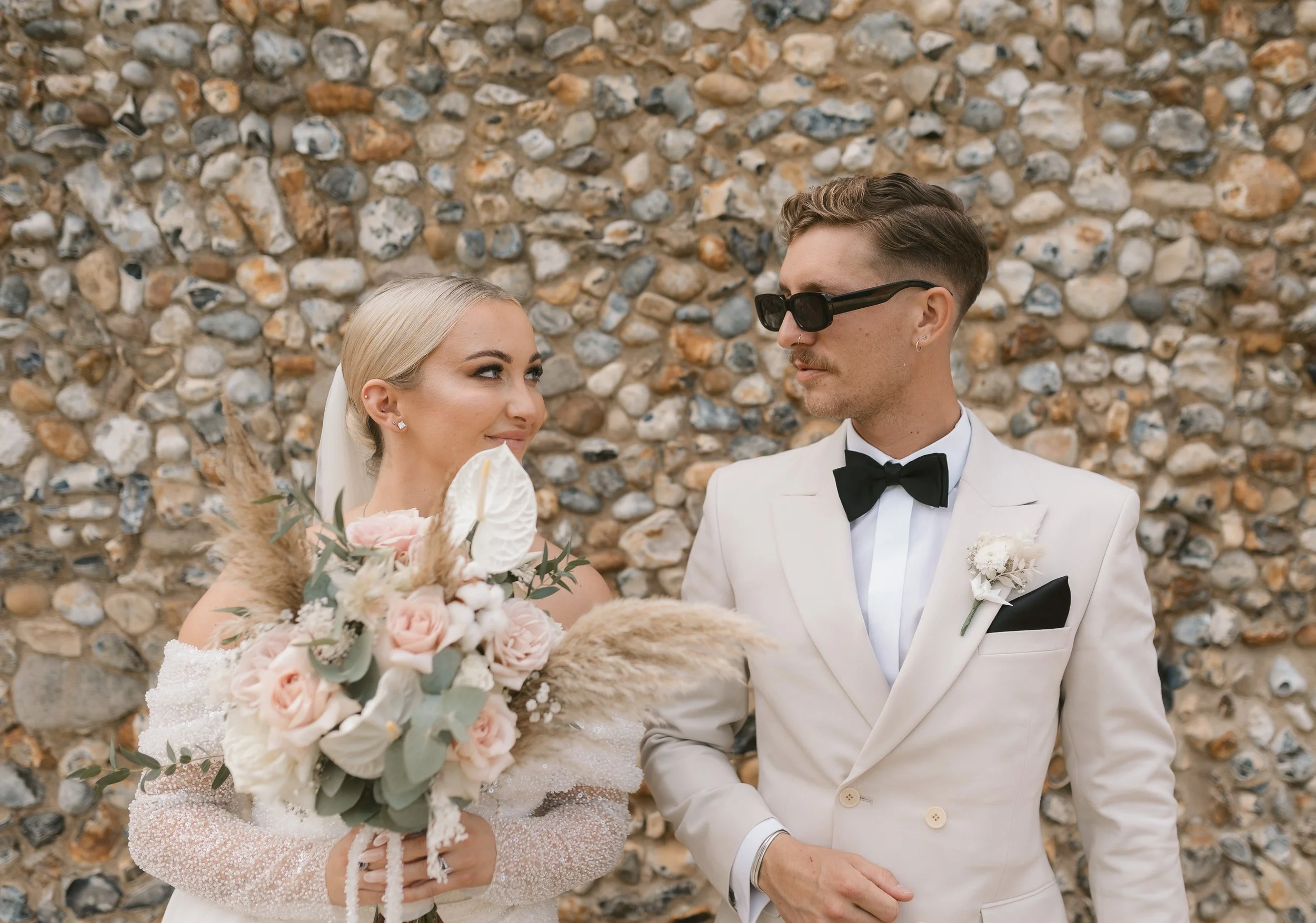 A bride and groom in wedding attire standing in front of a stone wall, gazing at each other. The bride holds a bouquet of pale pink and white flowers, and the groom wears sunglasses and a white tuxedo with a black bow tie.