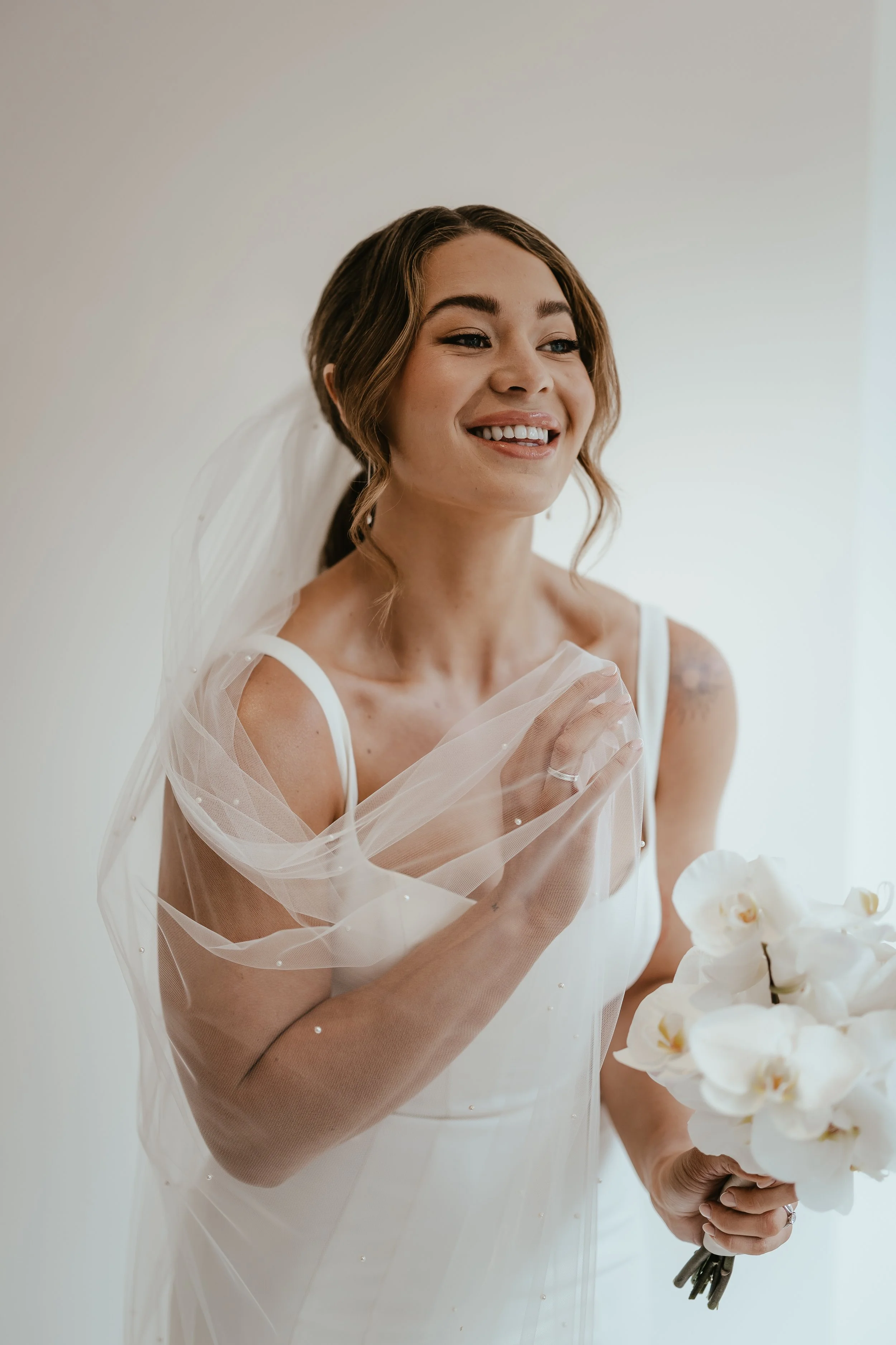 Bridal portrait of a smiling bride holding a bouquet of white orchids, wearing a white wedding dress and veil, with her hand on her chest, in a well-lit room.