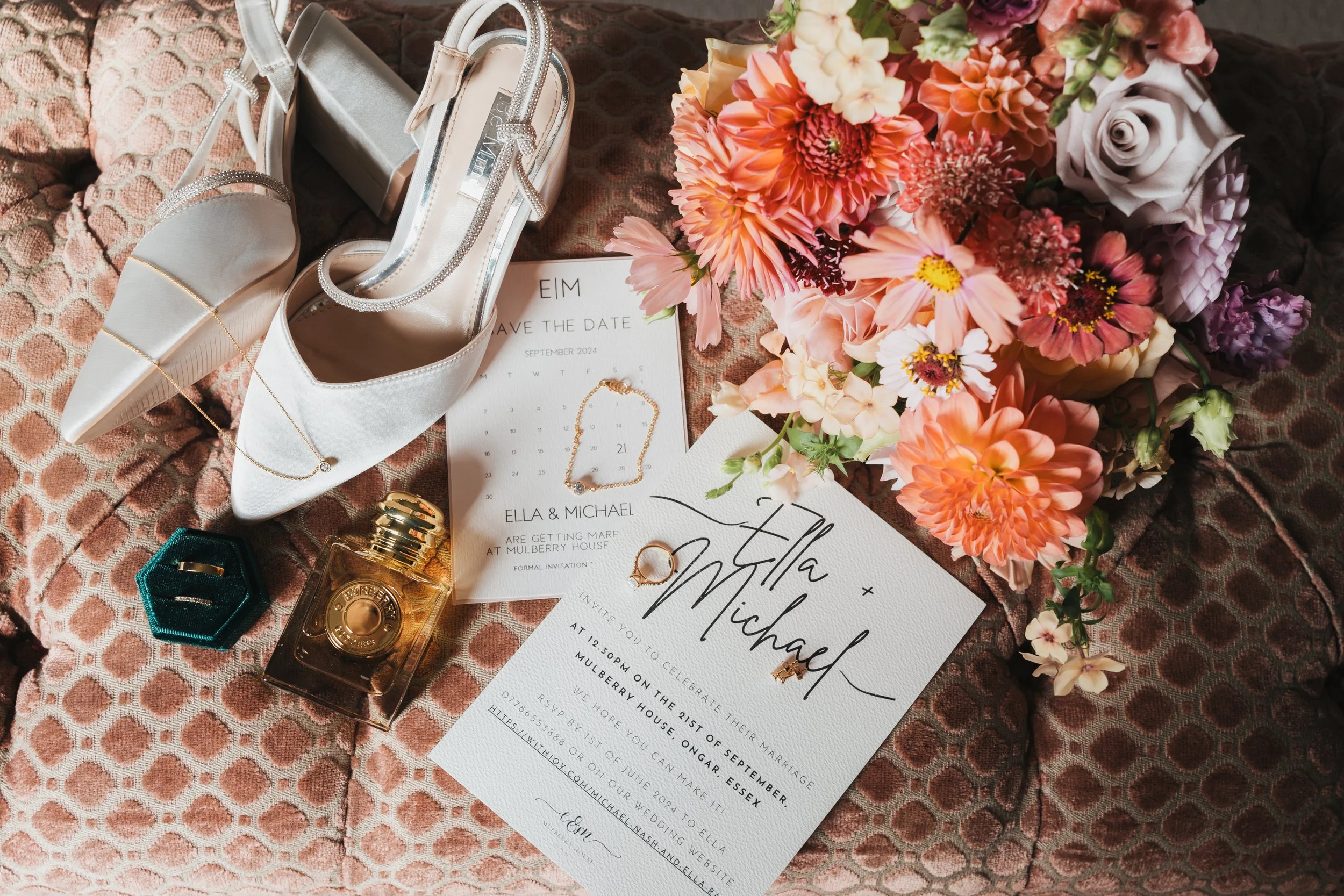 Wedding invitation, white high heels, perfume, jewelry, and a bouquet of pink and peach flowers on a patterned pink fabric surface.