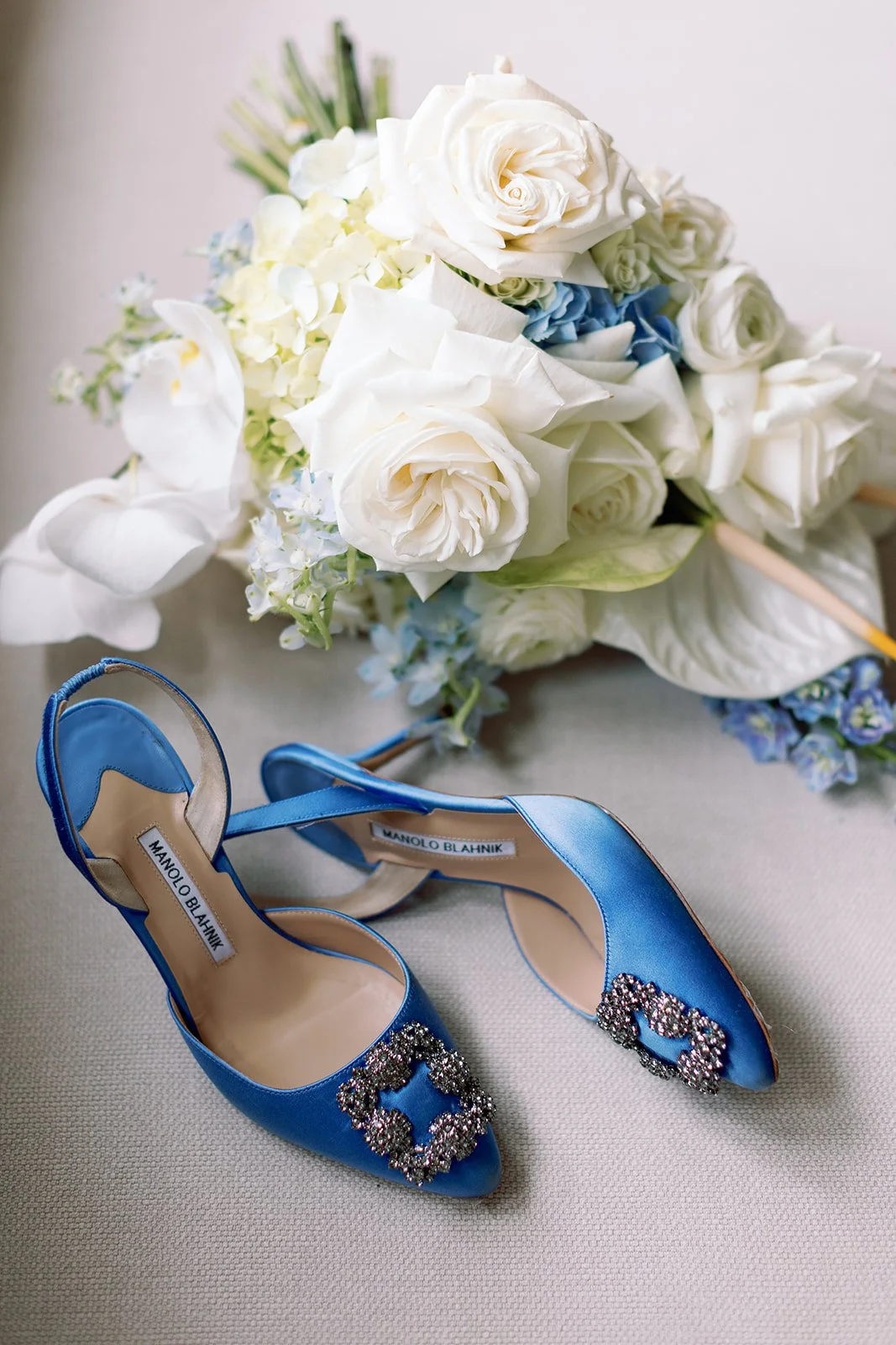 A pair of blue Manolo Blahnik high heels with jeweled embellishments on the toes, placed on a beige surface with a floral bouquet of white roses, white orchids, and blue flowers in the background.