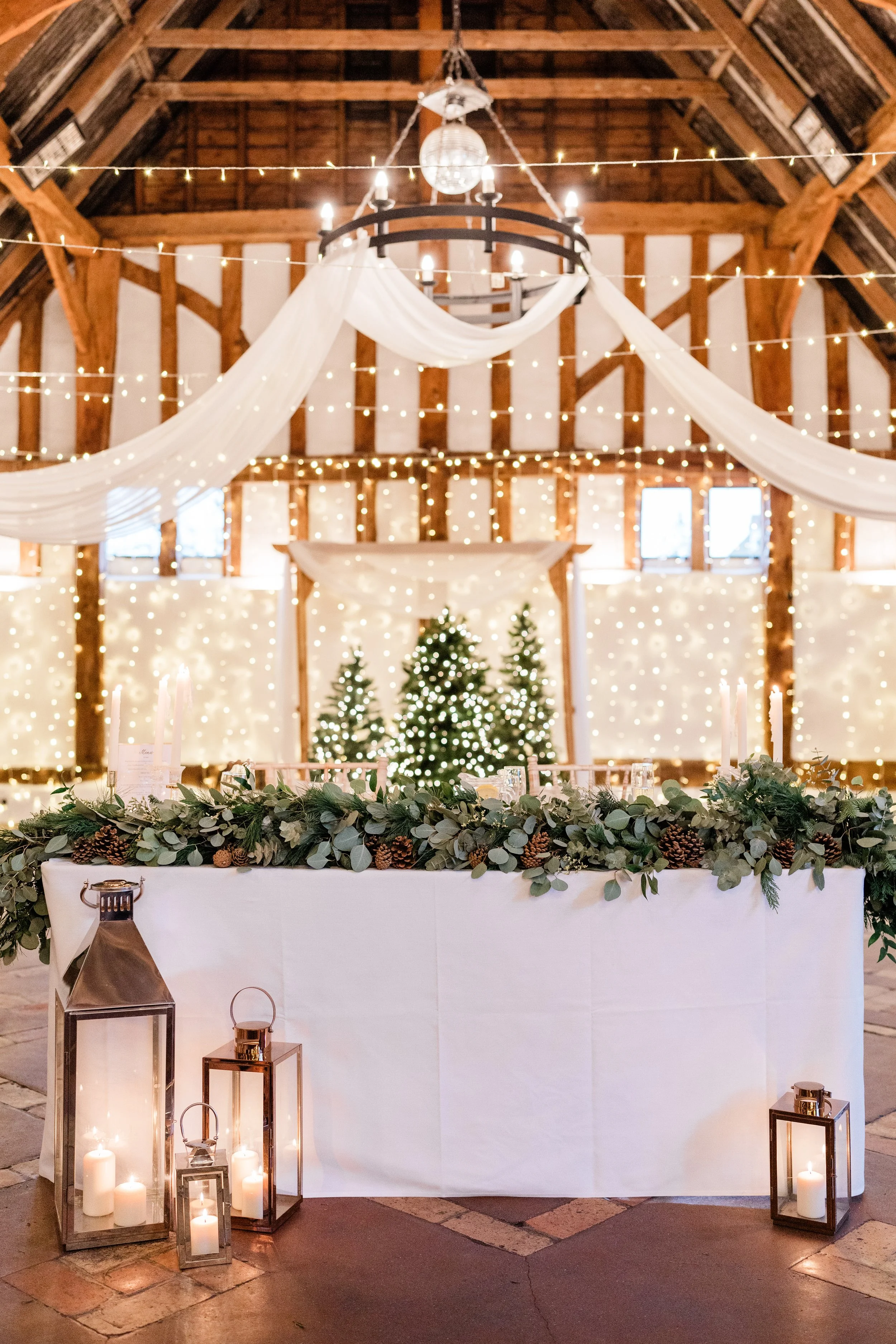 A festively decorated event space with a long table covered with a white cloth, decorated with greenery and pinecones. In the background, three decorated Christmas trees with white lights. The room has string lights, drapery, and a chandelier hanging