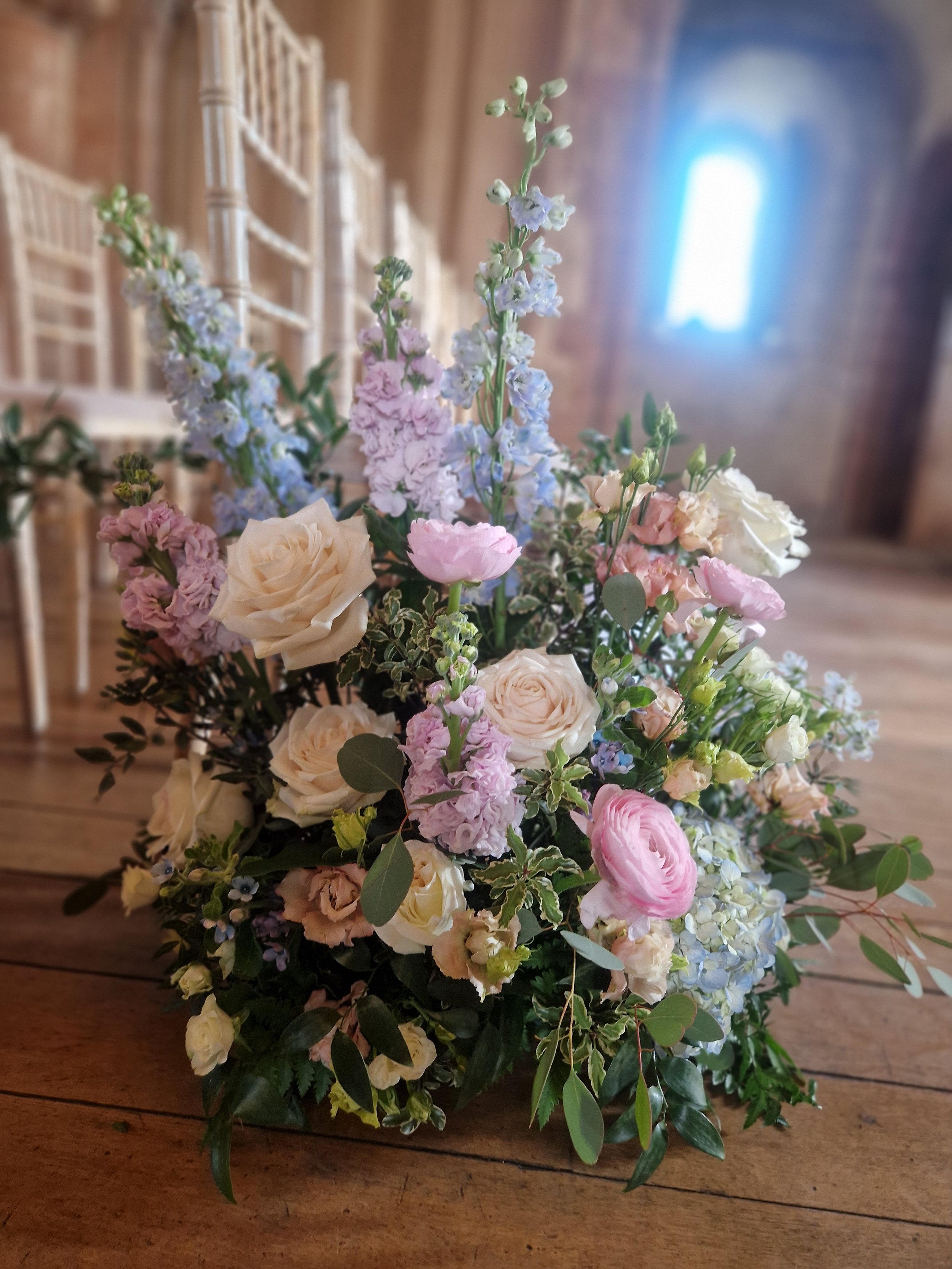 A bouquet of pastel-colored flowers including roses, delphiniums, and ranunculus on a wooden table inside a rustic brick building with chairs in the background.