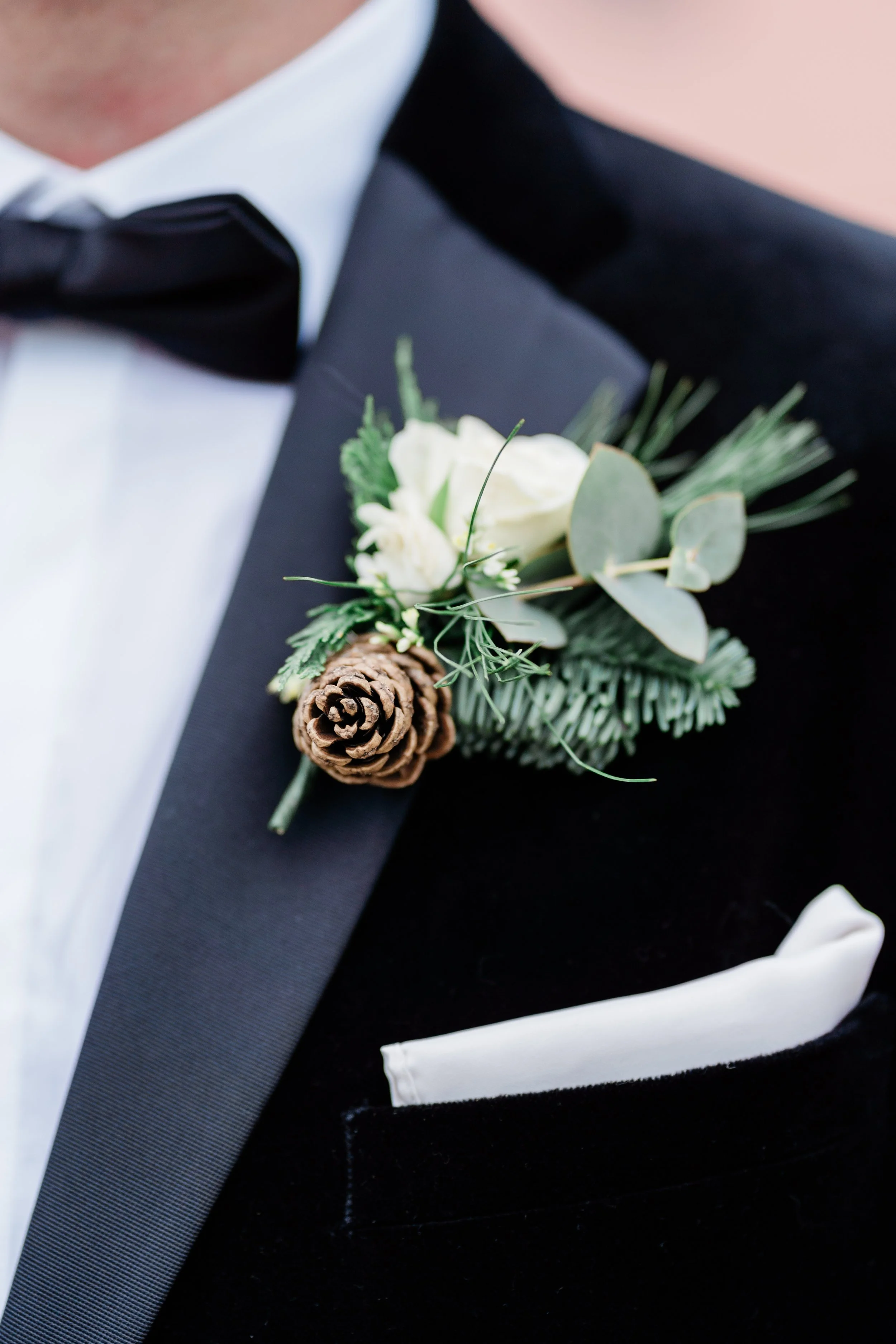 Close-up of a man's tuxedo with a boutonniere featuring a white rose, pine cone, and greenery.