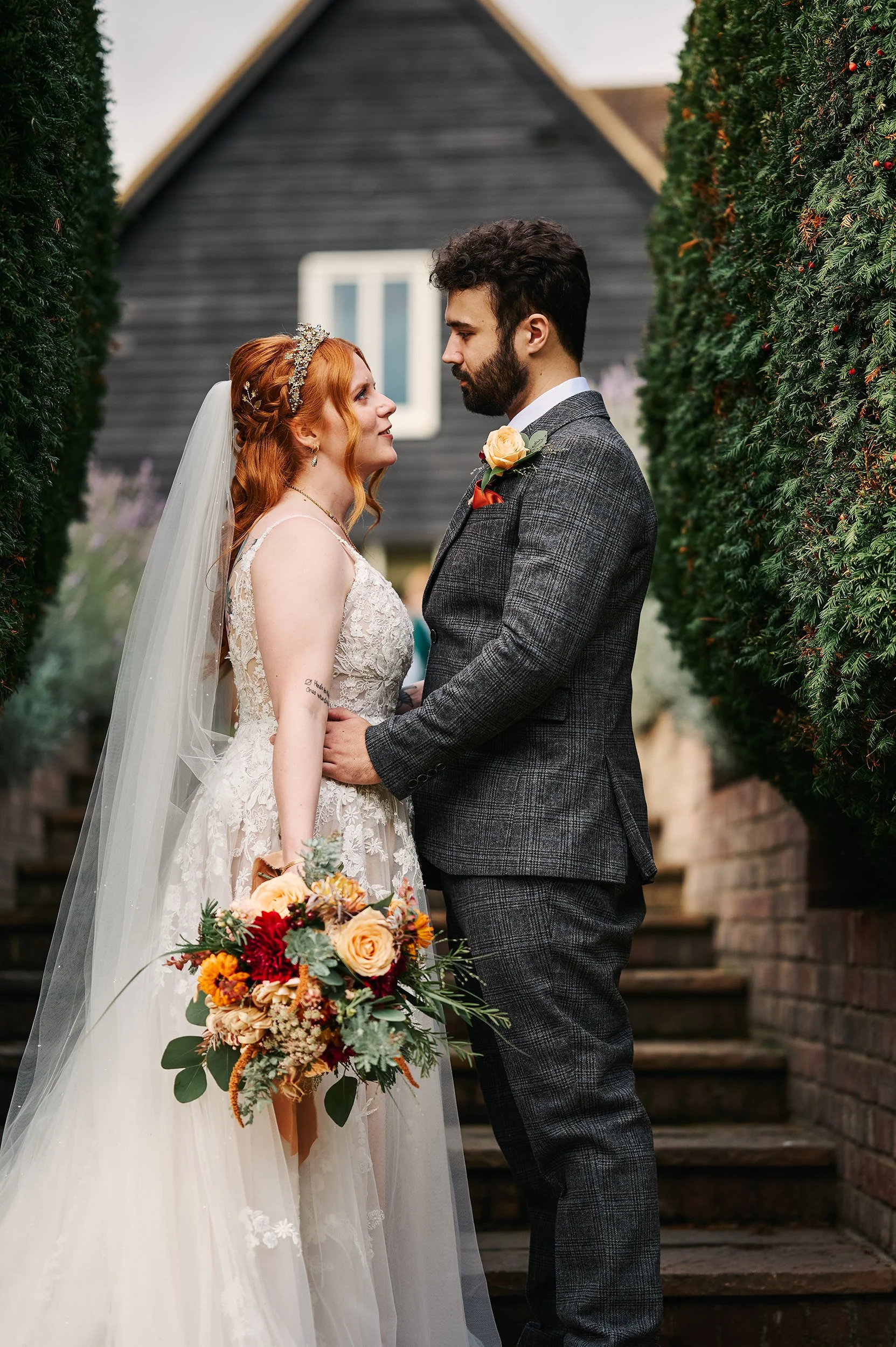 A bride and groom stand close together outdoors, gazing into each other's eyes. The bride has long red hair, wears a white wedding gown with lace details, a veil, and holds a colorful bouquet. The groom has dark hair and a beard, dressed in a gray ch
