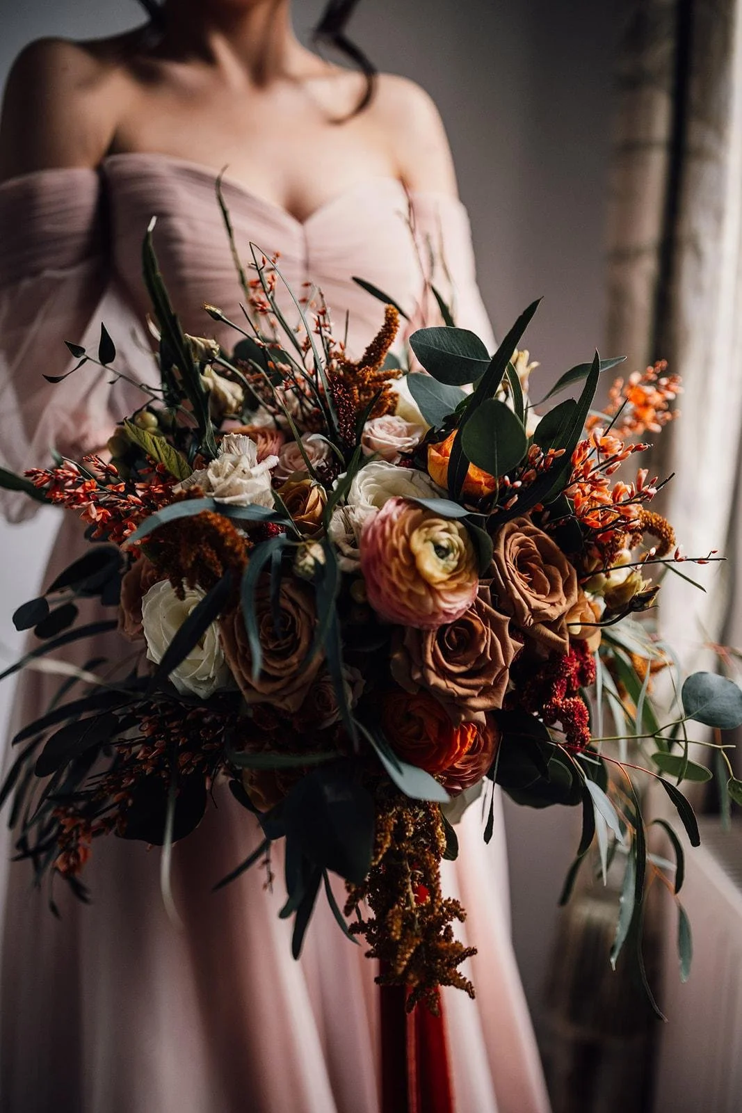 A woman in a pink off-the-shoulder dress holding a large, colorful bouquet of flowers with roses, ranunculus, and greenery, standing near a window.