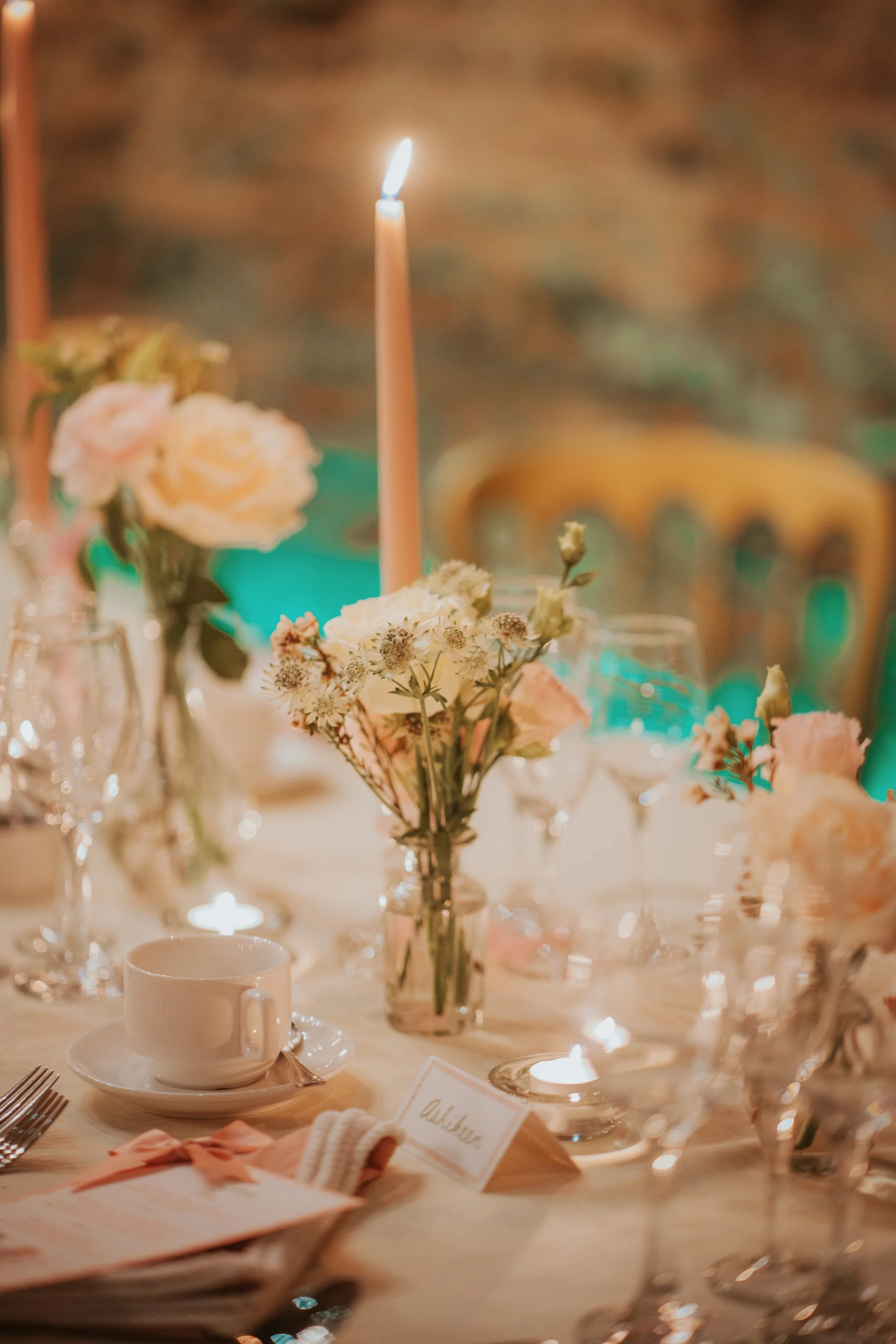 A decorated dining table with pink and white flowers, a lit pink candle, wine glasses, a white cup and saucer, and a place card, indicating a special occasion or event.