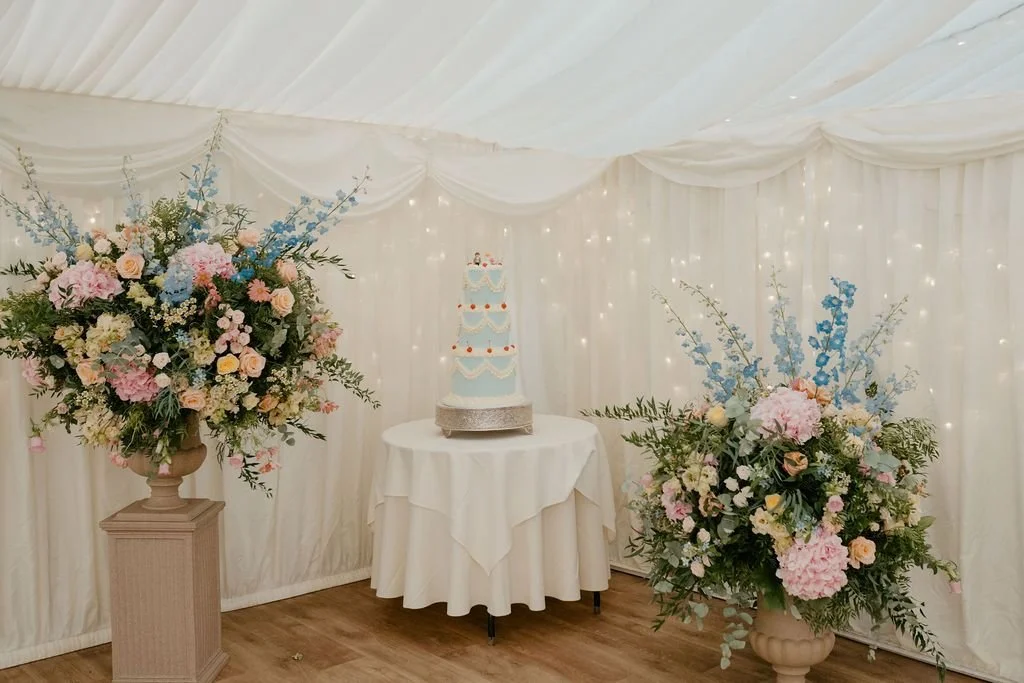 A wedding cake on a round table with a white tablecloth, two large floral arrangements on each side, and a backdrop of draped white fabric with string lights.
