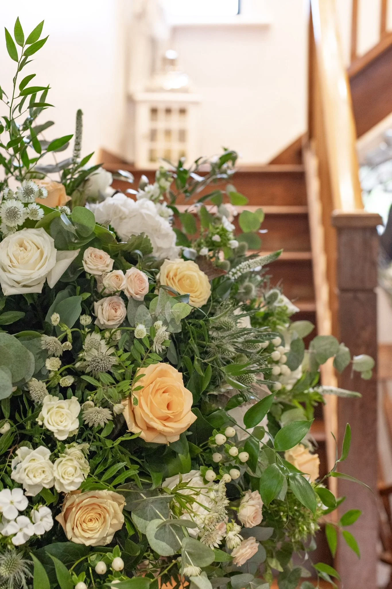 Close-up of a floral arrangement with cream and peach roses, white and green foliage, and small white berries, placed near wooden stairs in an indoor setting.