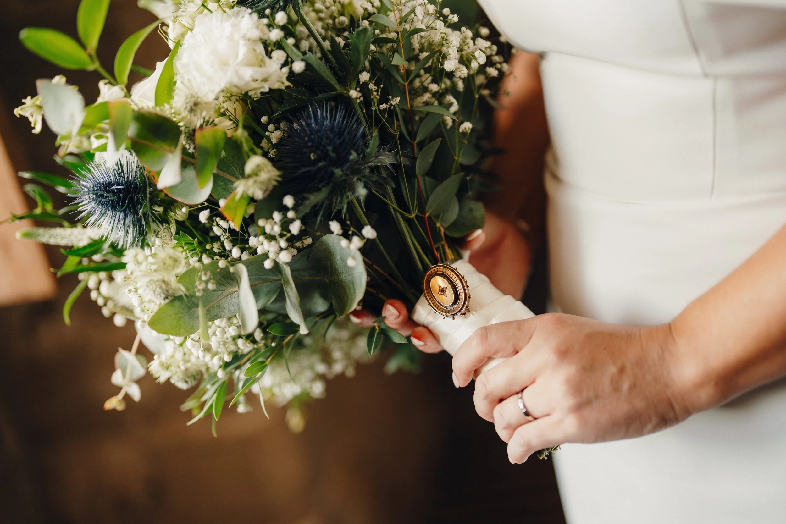 A bride holding a bouquet of white and dark flowers, including white baby's breath and dark thistles, with her left hand showing a wedding ring.