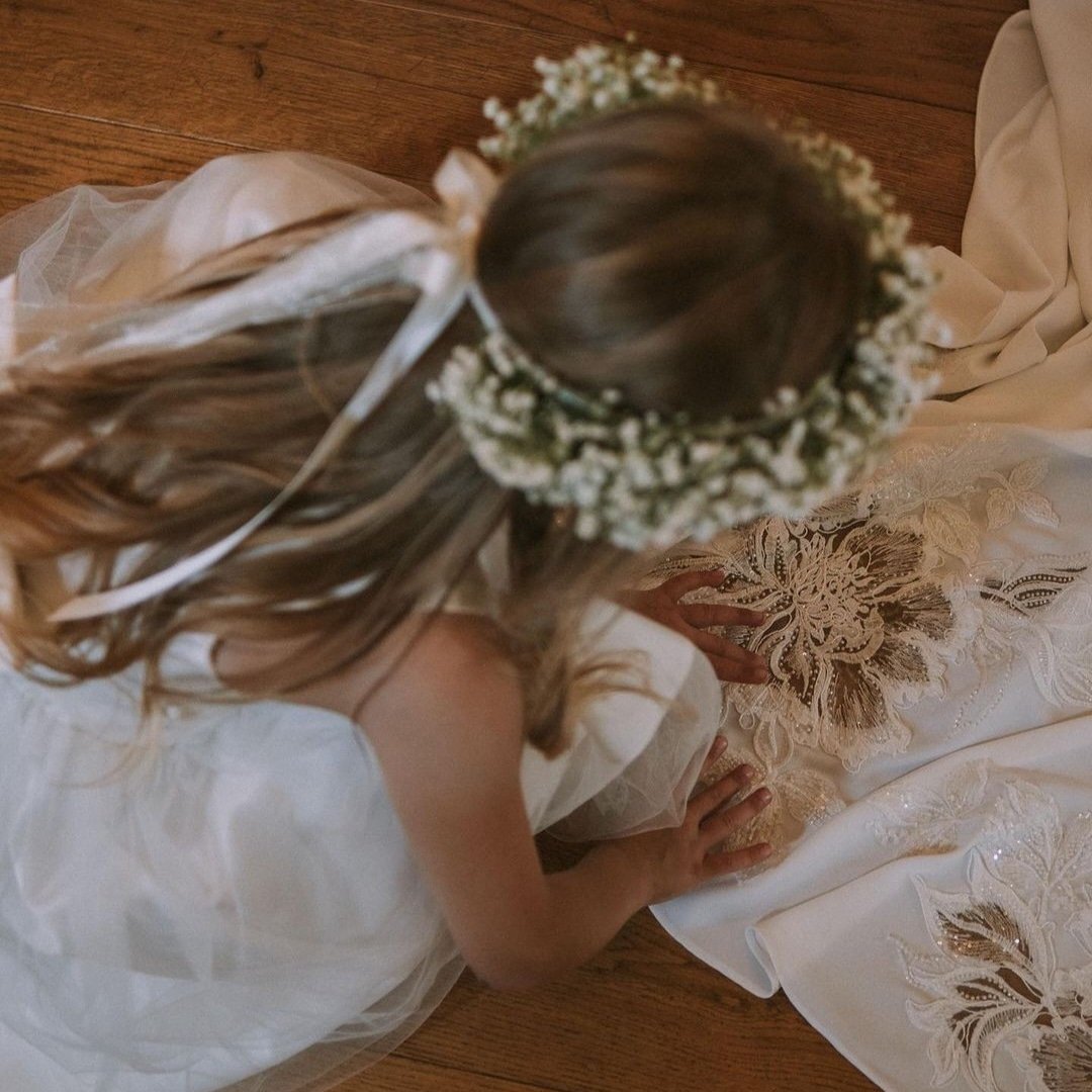 A young girl with long wavy hair wearing a white dress and a floral crown, kneeling on a wooden floor, gently touching a piece of embroidered fabric.