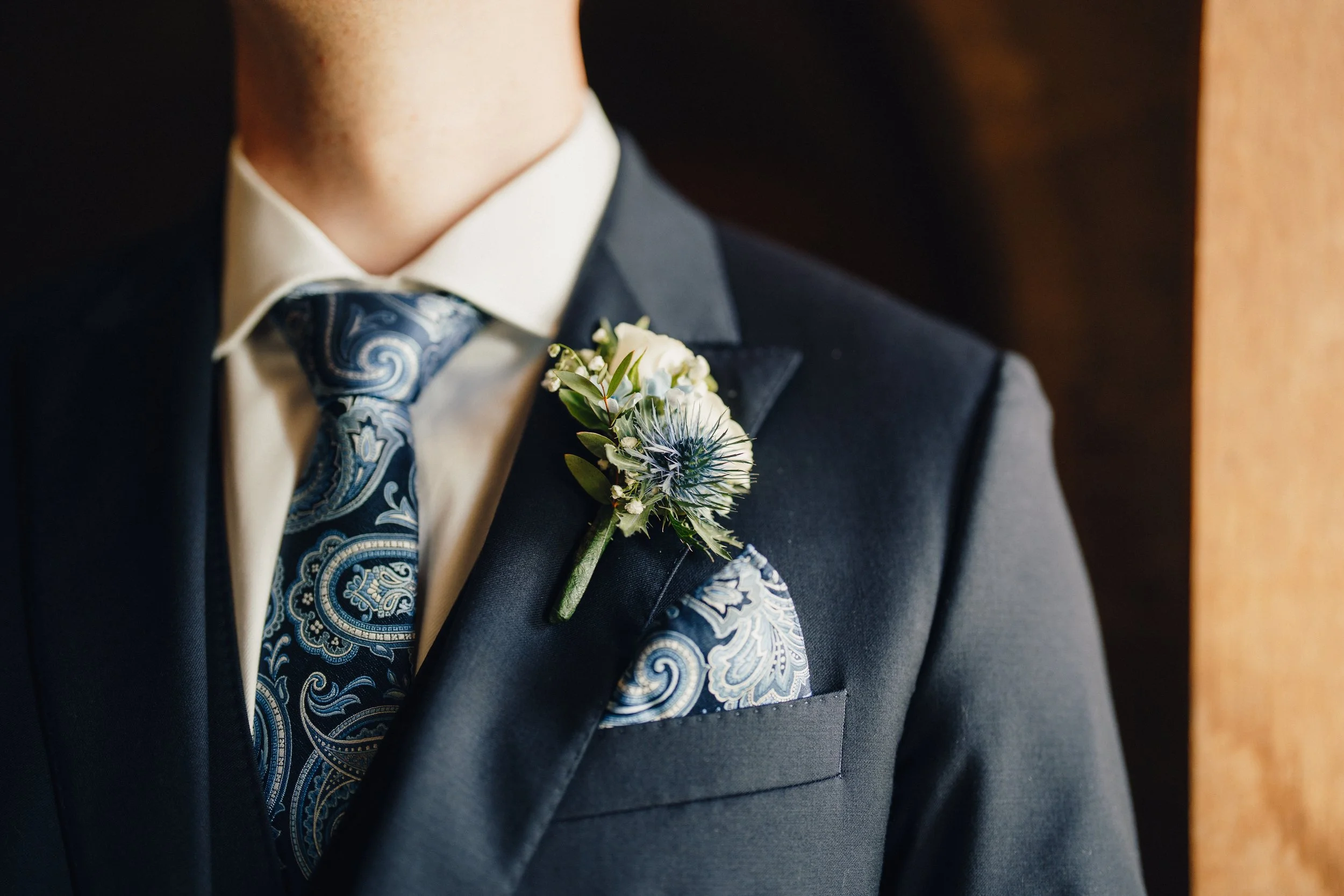 Close-up of a groom wearing a dark suit, paisley tie, white shirt, with a boutonniere and matching pocket square.