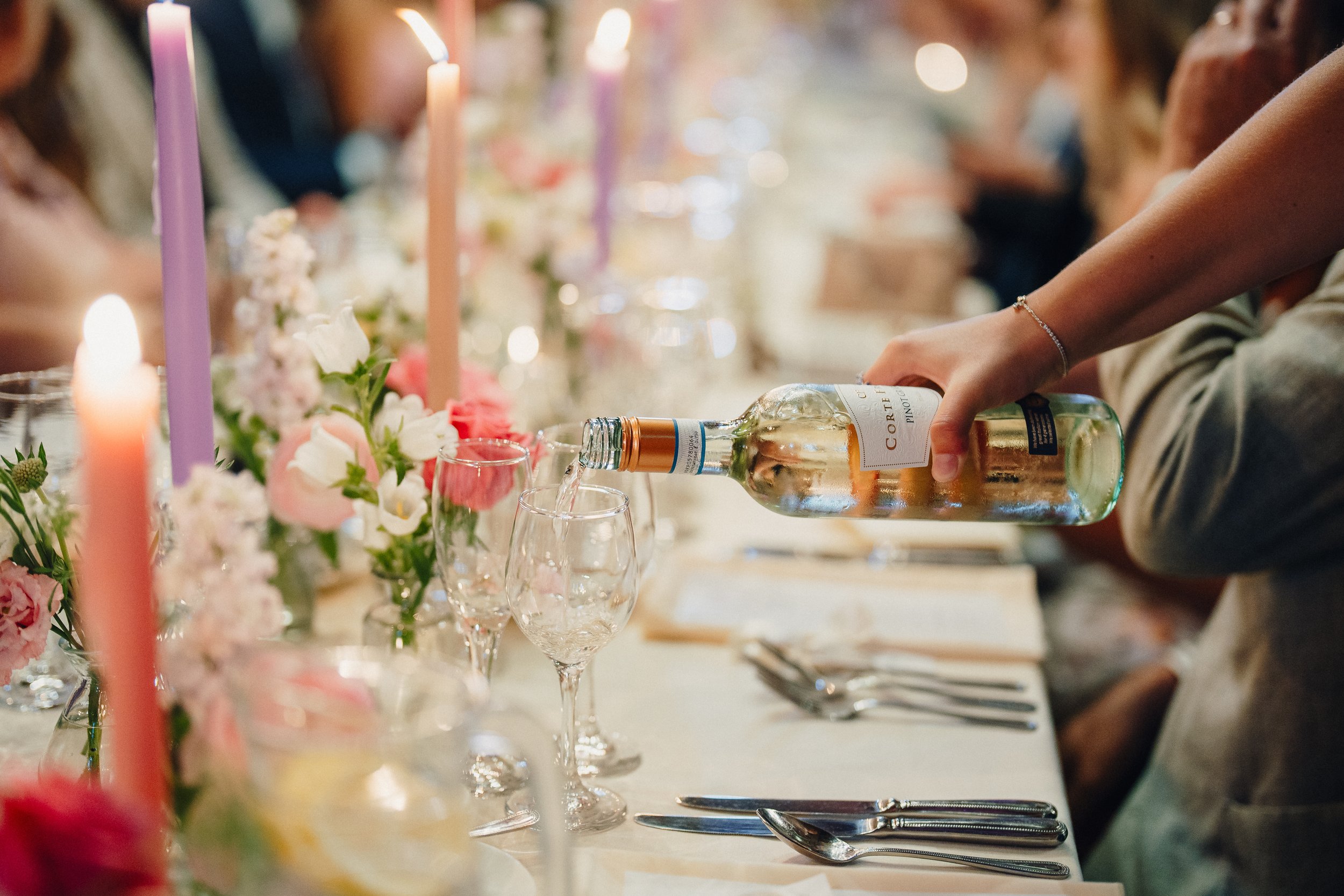 Person pouring wine into a glass at a decorated dinner party with floral centerpieces and candles.
