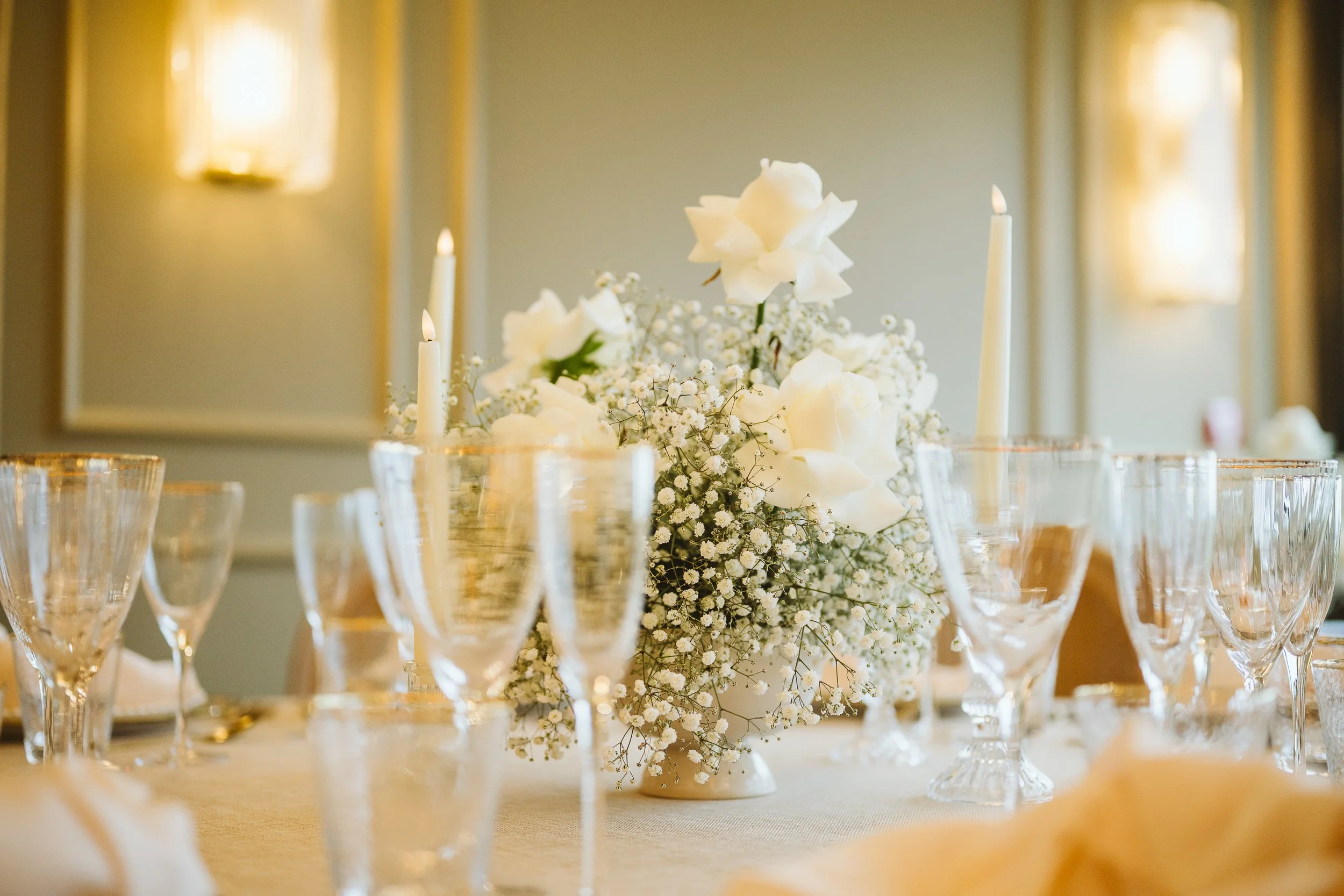 Elegant wedding table setting with a floral centerpiece of white roses and baby's breath, surrounded by tall lit white candles and empty champagne glasses.