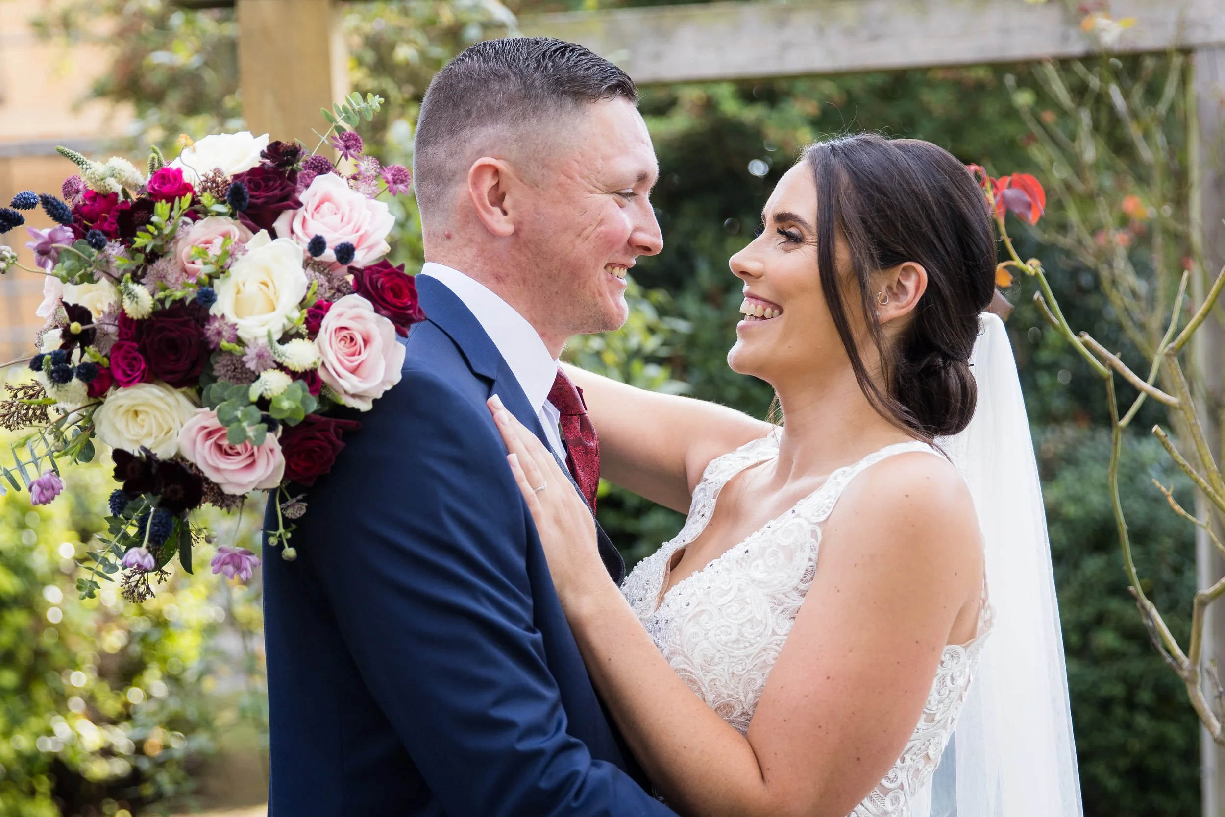 A bride and groom smiling at each other outdoors, with the bride holding a large colorful bouquet of flowers.