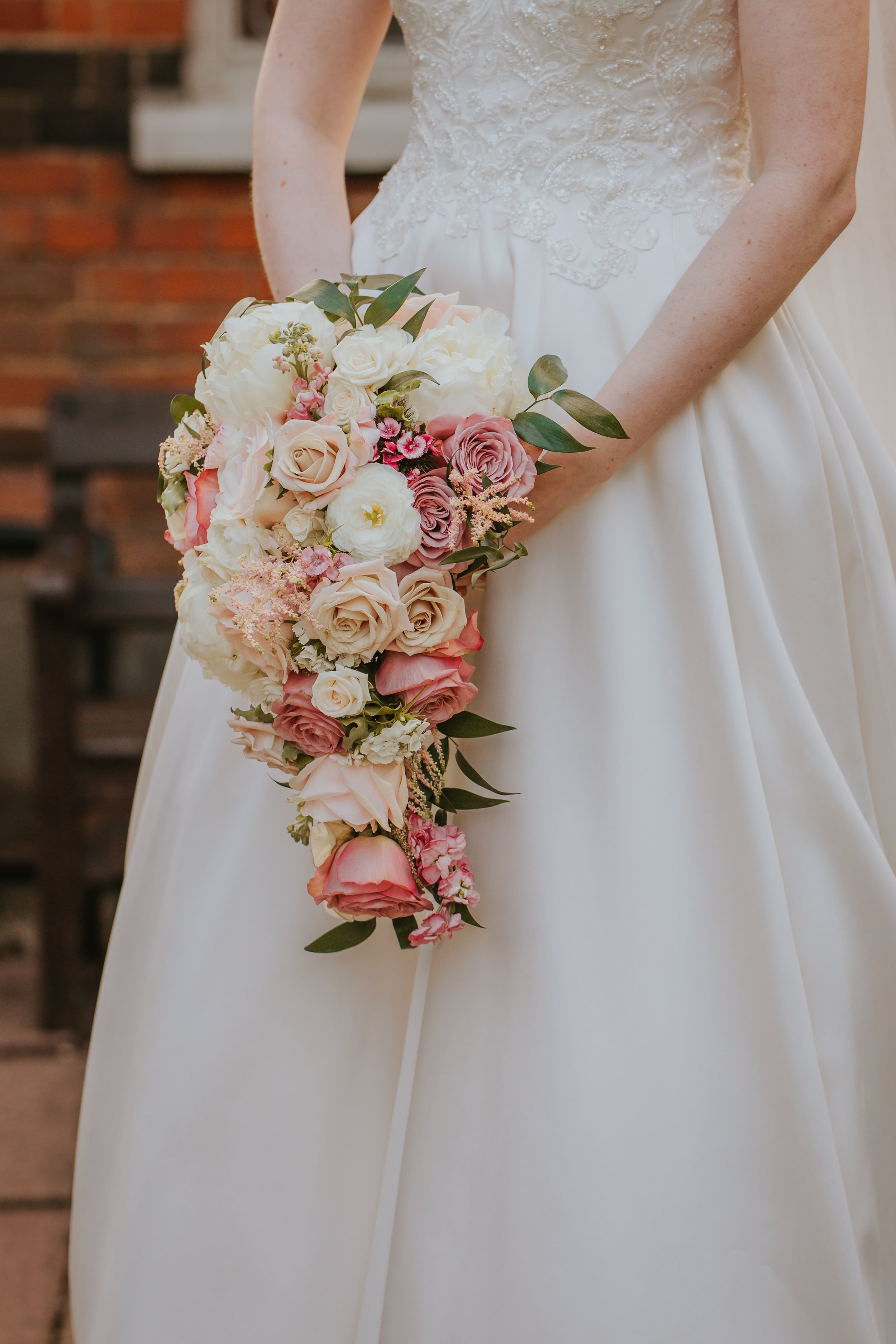 Close-up of a bride holding a cascading bouquet of pink, white, and cream flowers in front of her white wedding dress with lace detail.