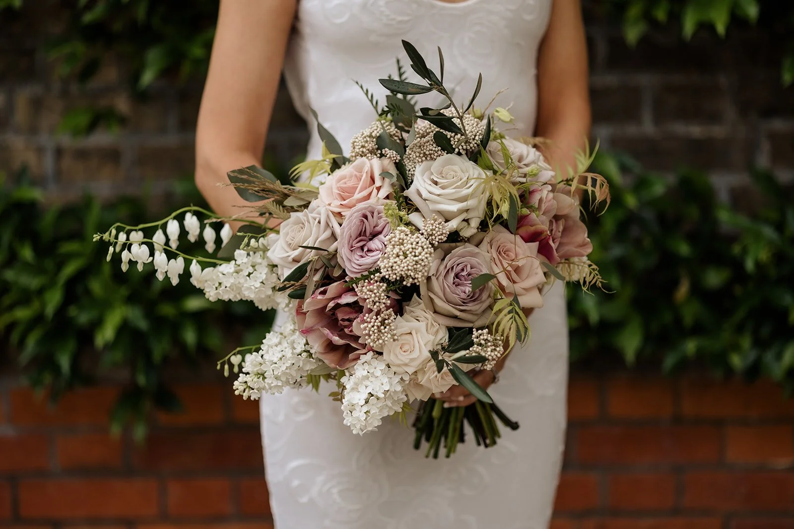 A woman in a white dress holding a large bouquet of white, blush, and purple flowers with greenery against a brick wall and green foliage background.