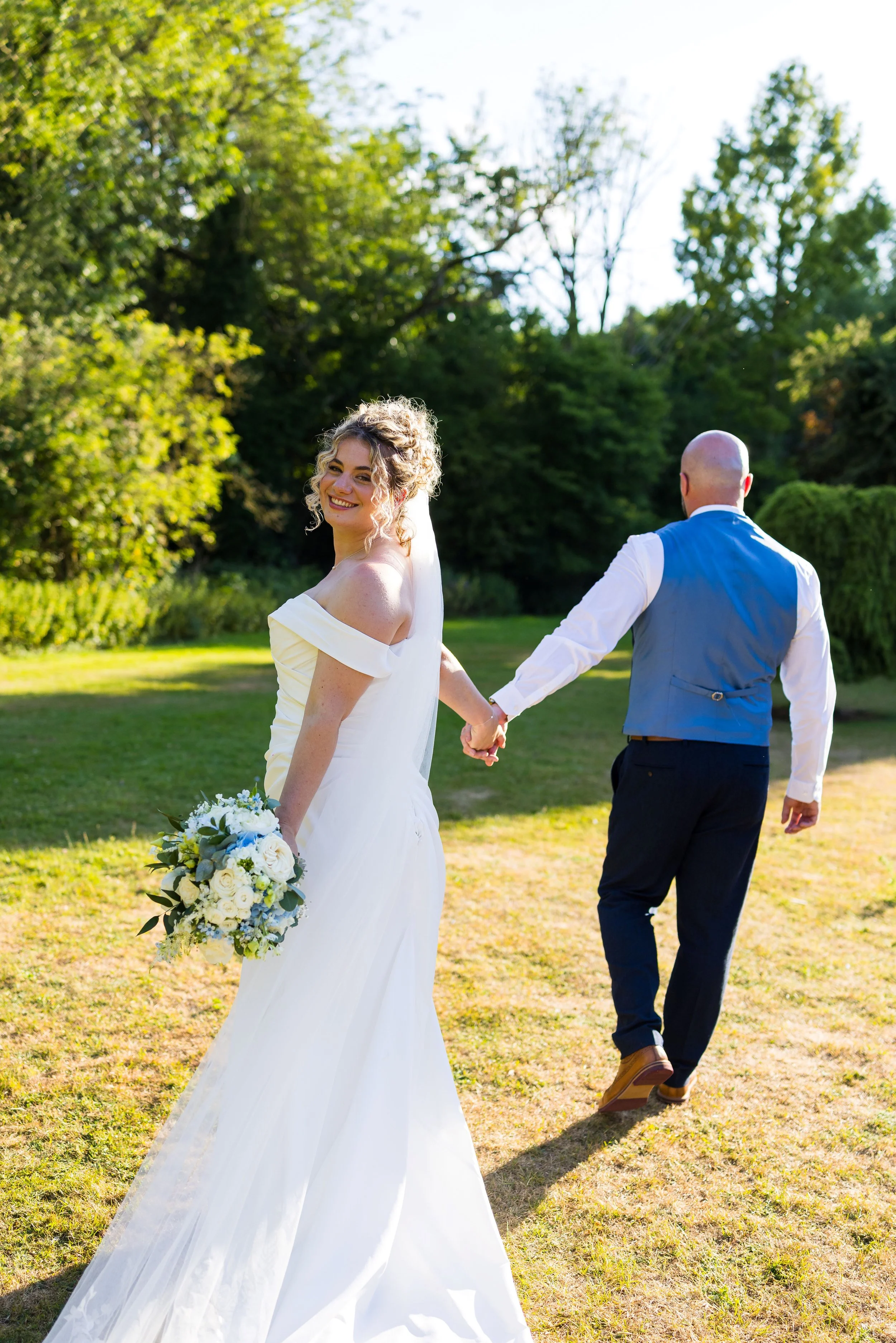 A smiling bride in a white wedding dress holding a bouquet of white and blue flowers, looking back over her shoulder while holding hands with her groom, dressed in a white shirt, blue vest, and dark pants, walking away on a grassy field during a sunn