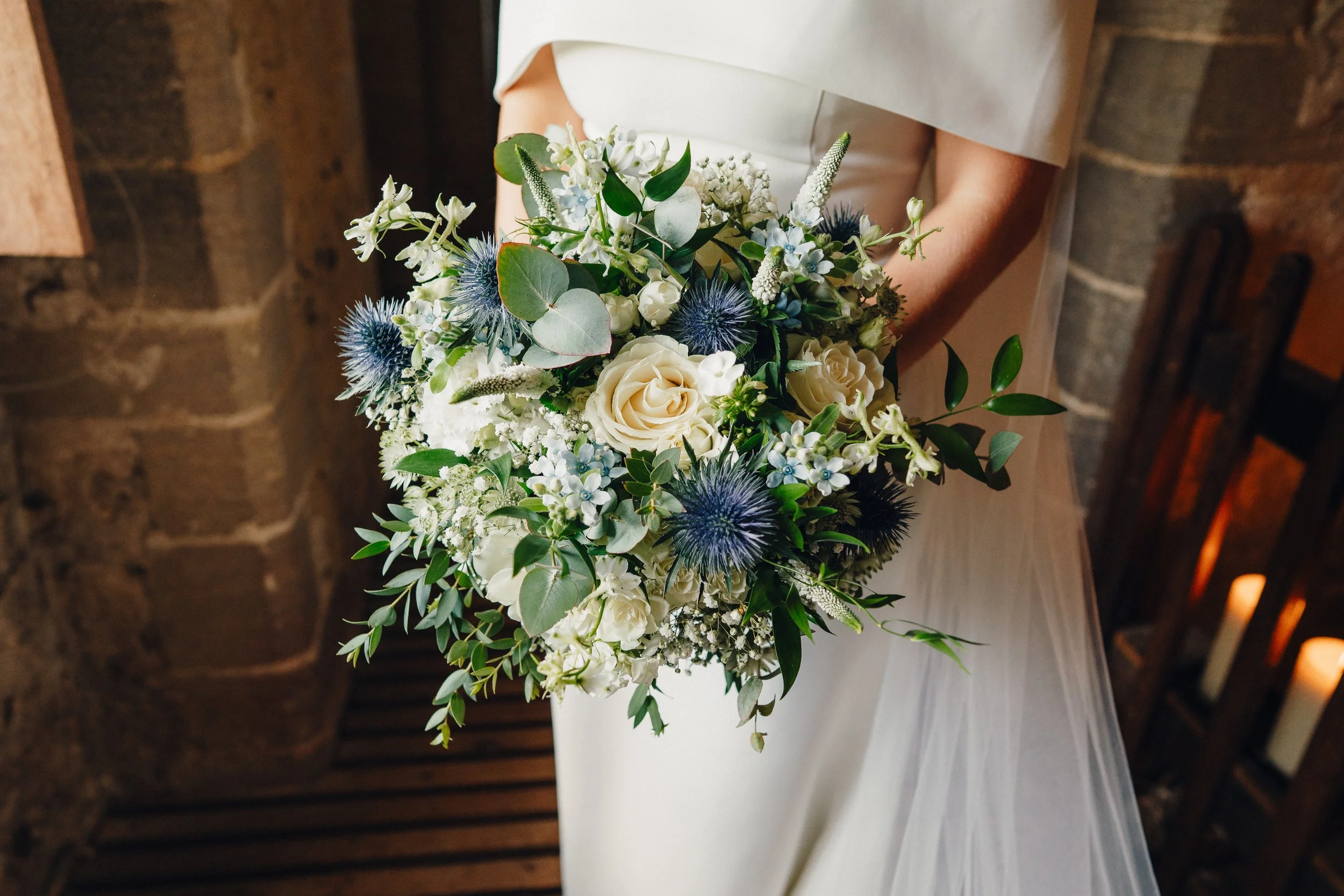 Bride holding a large bouquet of white roses, blue thistles, and greenery, in a cream wedding dress inside a rustic venue.