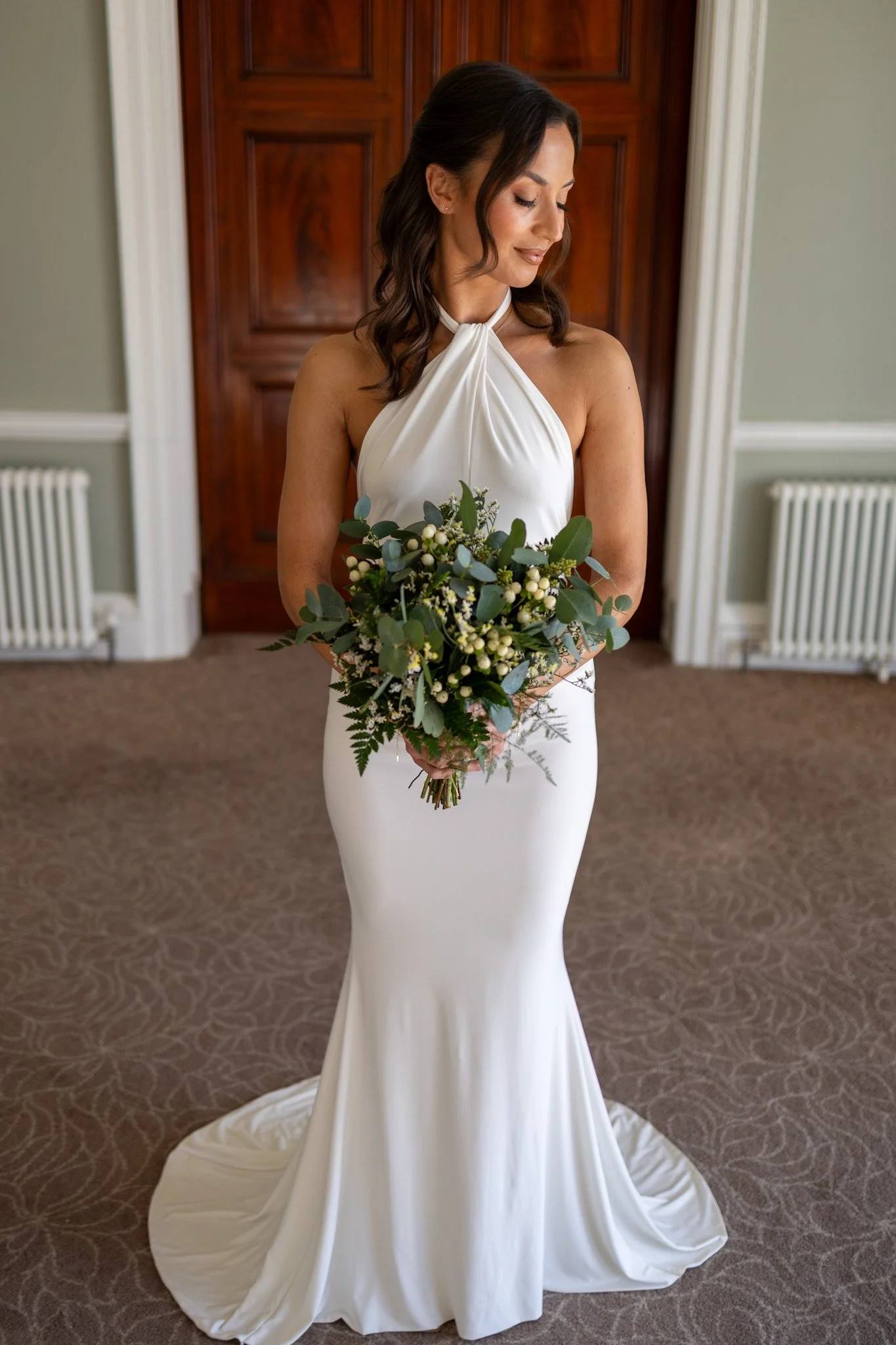 A bride in a white wedding dress holding a bouquet of greenery and white flowers, standing in a room with green walls, white trim, and wooden doors.