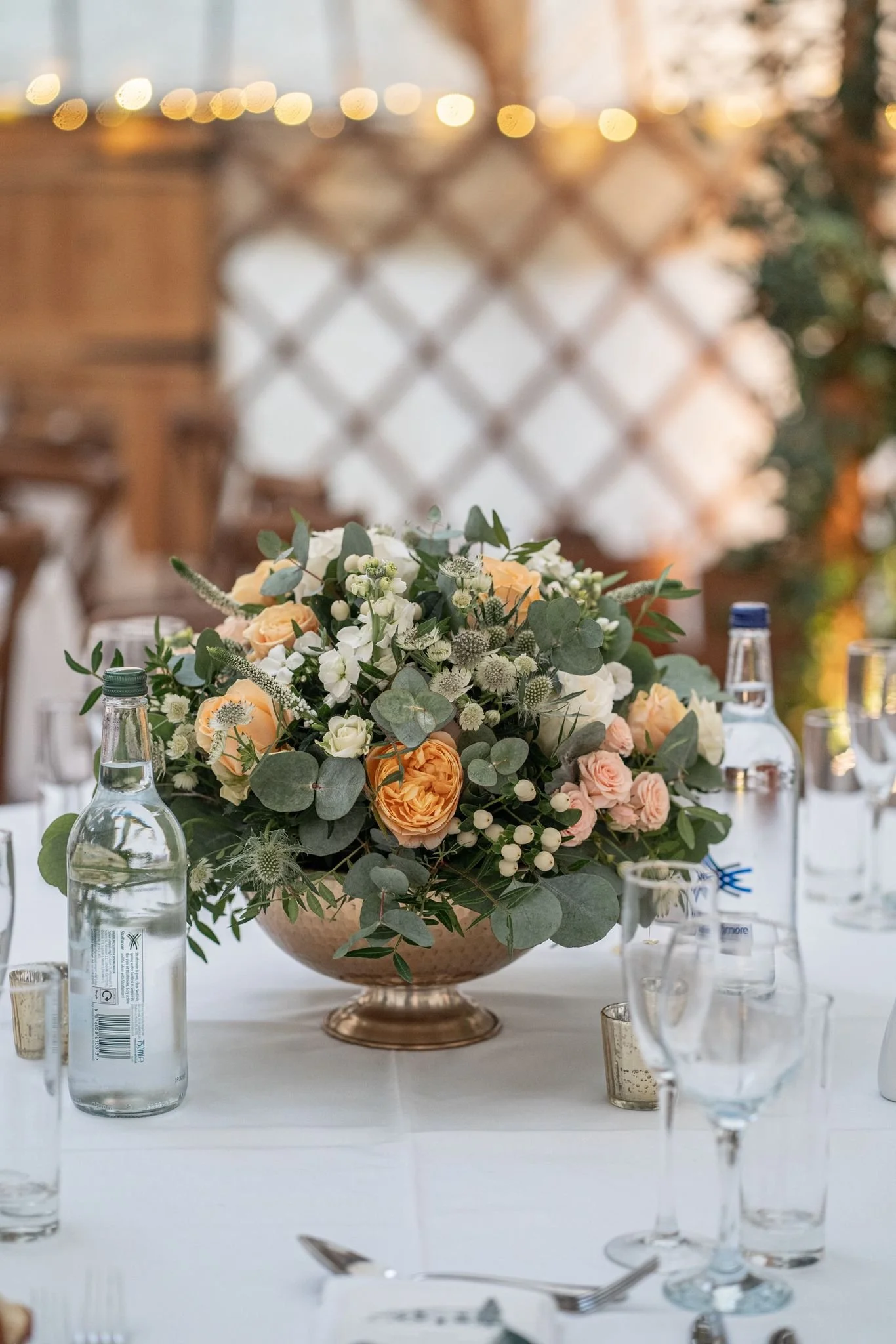 A floral centerpiece with peach, white, and blush roses, white stock, eucalyptus, and other greenery on a banquet table at an outdoor event with string lights in the background.