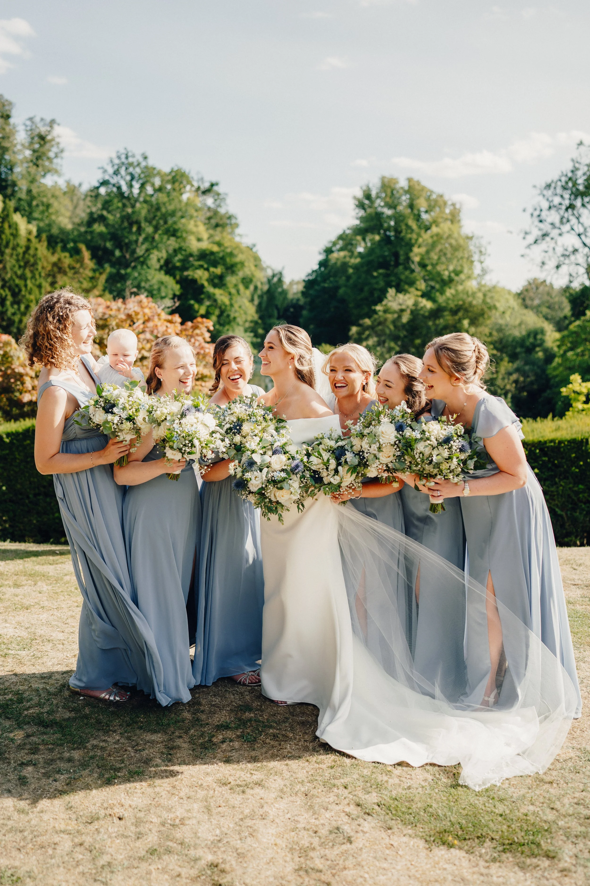 A bride in a white wedding dress surrounded by her bridesmaids in matching light blue dresses, all holding bouquets, outdoors on a sunny day with trees in the background.