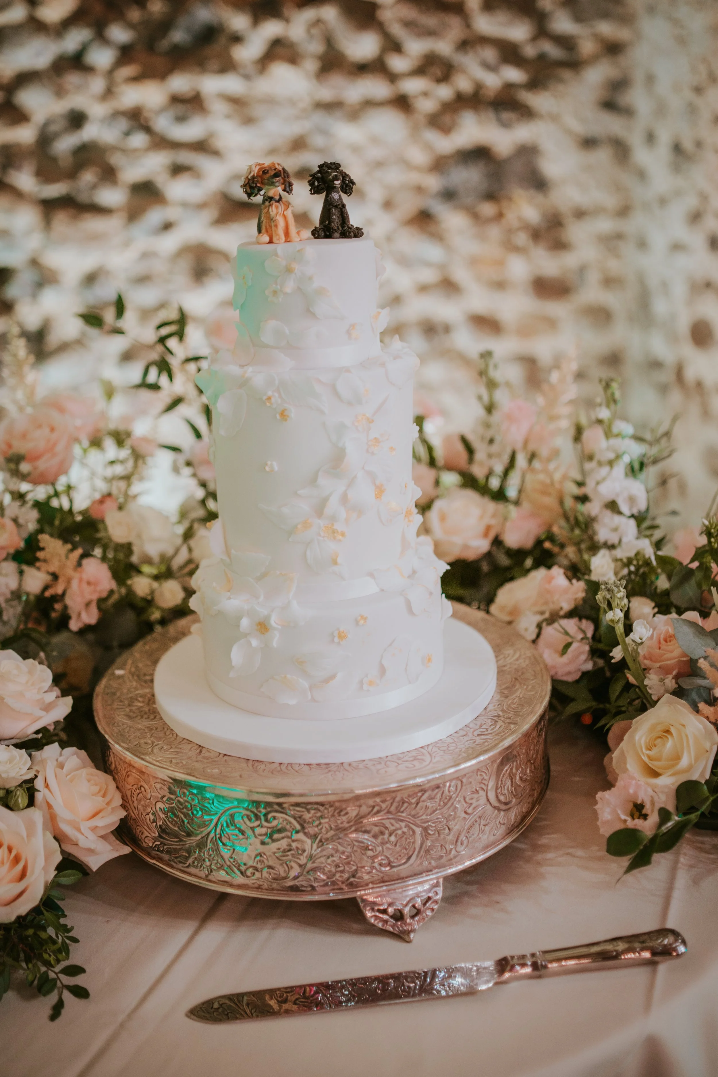 Elegant three-tiered white wedding cake with floral decorations, topped with two small dog figurines, surrounded by pink and white roses, on a decorative silver cake stand.