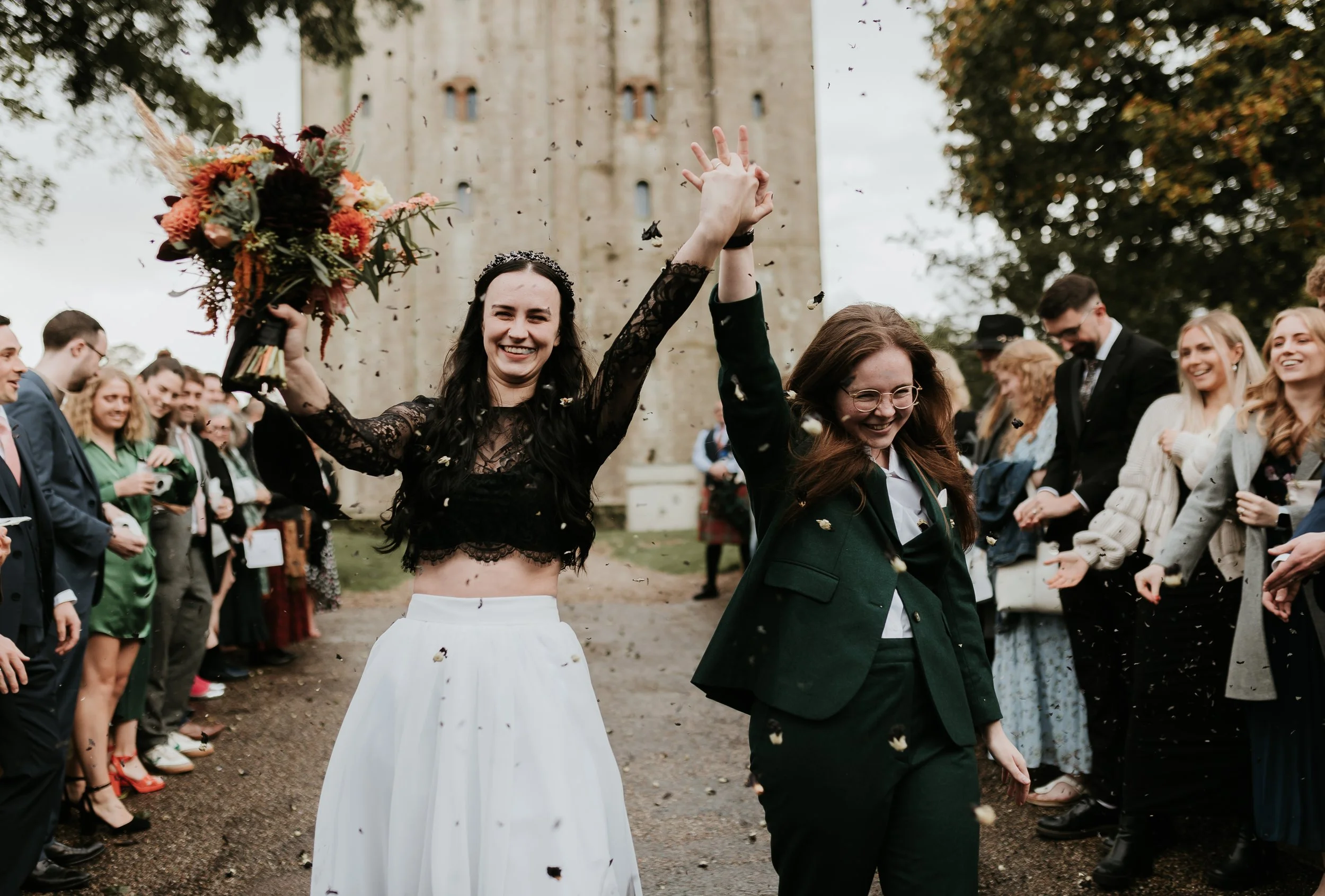 Two women, one in a wedding dress and another in a green suit, celebrating outdoors with a crowd, throwing confetti, in front of a stone tower.