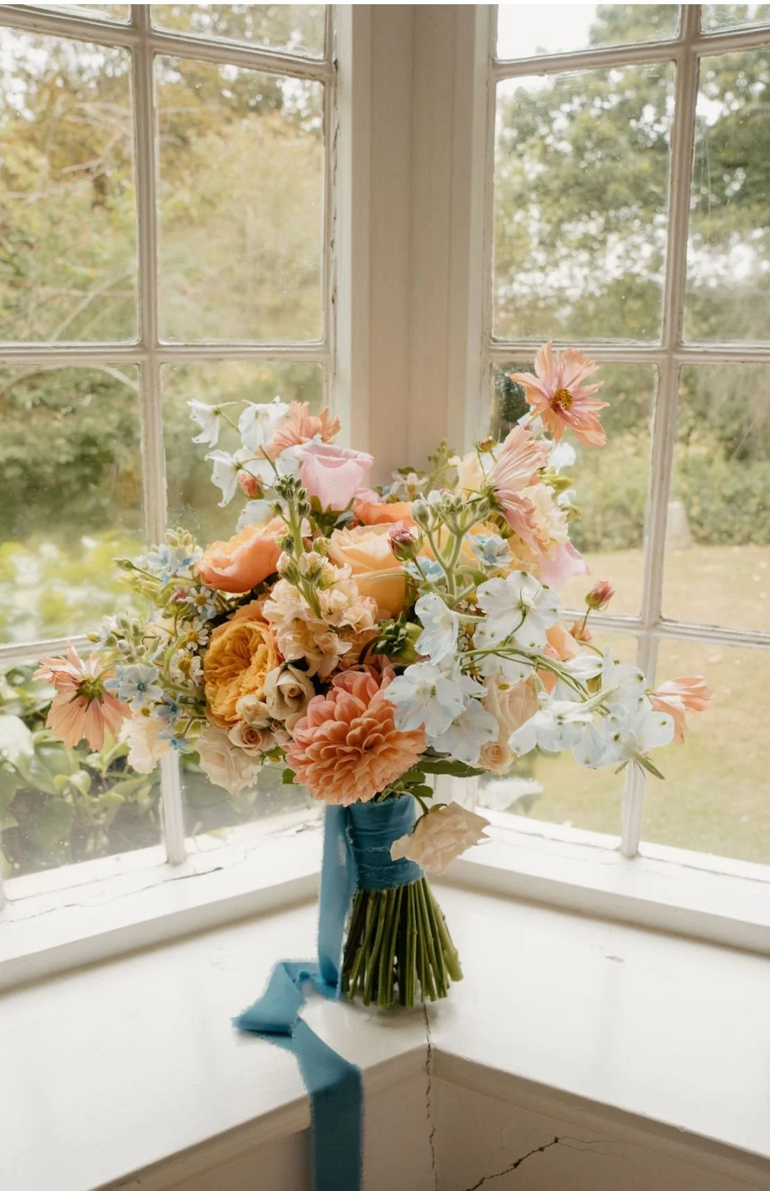 A bouquet of pastel-colored flowers in a blue ribbon-wrapped vase, placed on a white windowsill with a multi-pane window showing a blurred outdoor garden scene.
