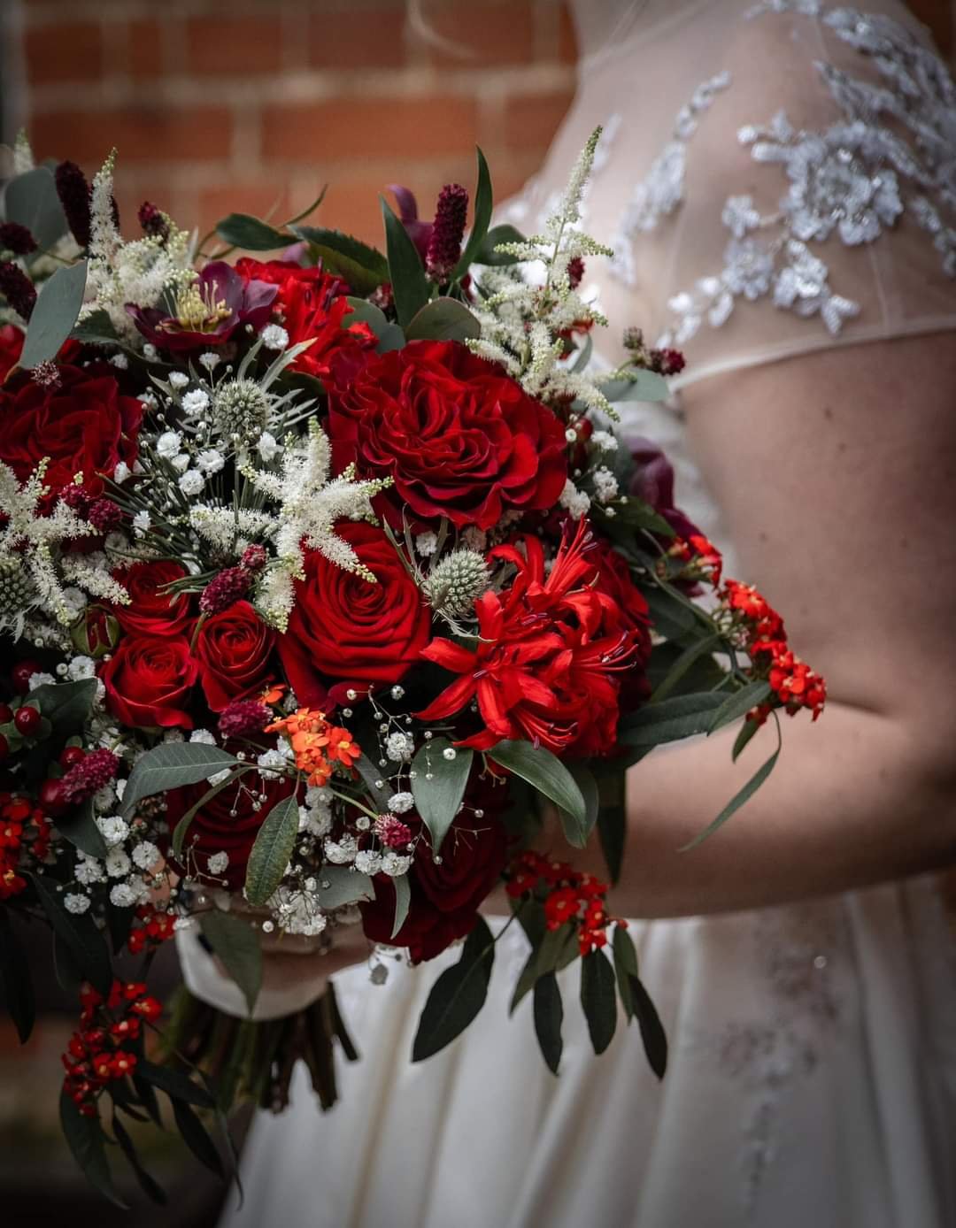 Close-up of a bride holding a bouquet of red roses, white baby's breath, and greenery, with a lace wedding dress visible.