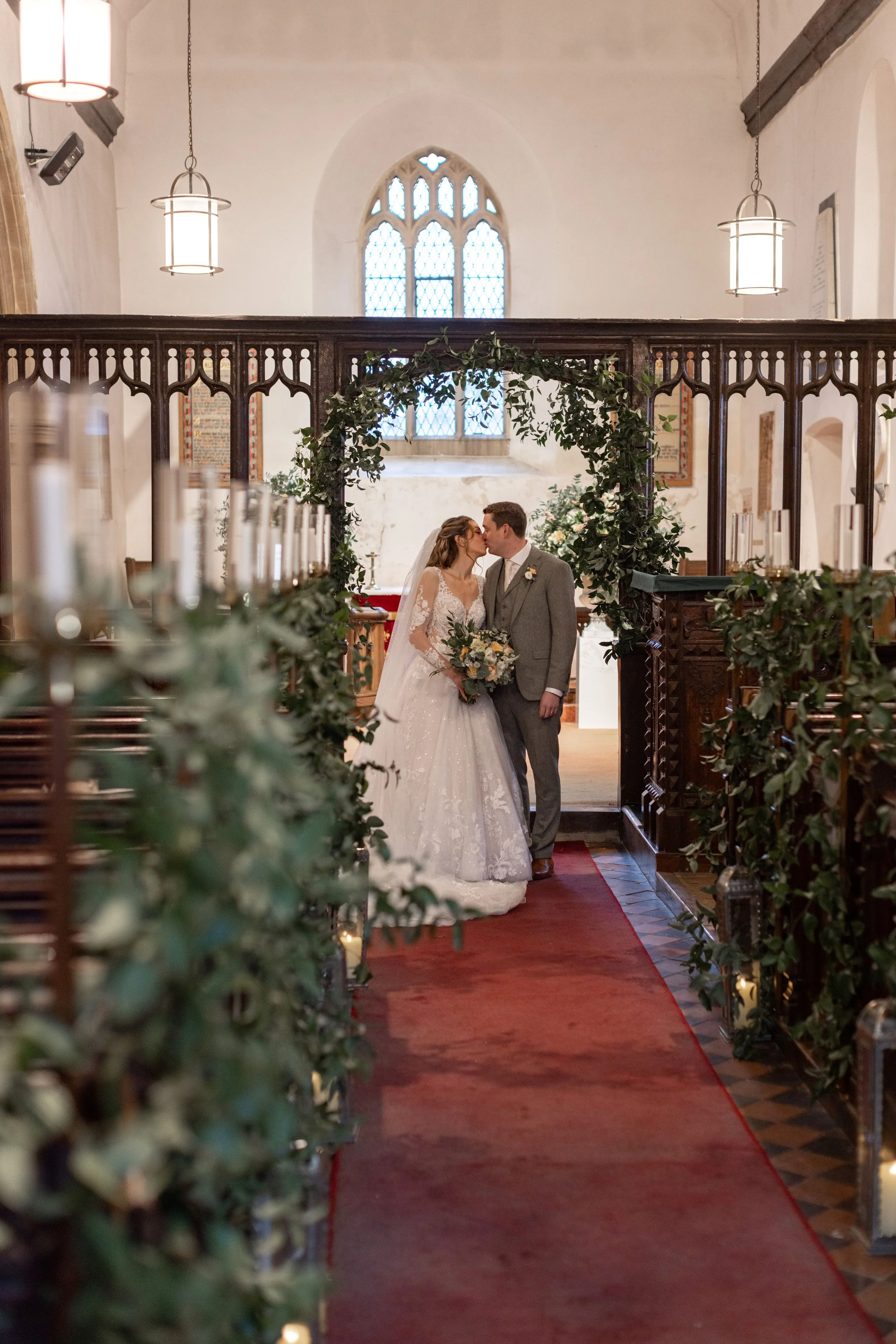 A bride and groom sharing a kiss at their wedding ceremony inside a church, decorated with greenery and candles, with stained glass windows in the background.