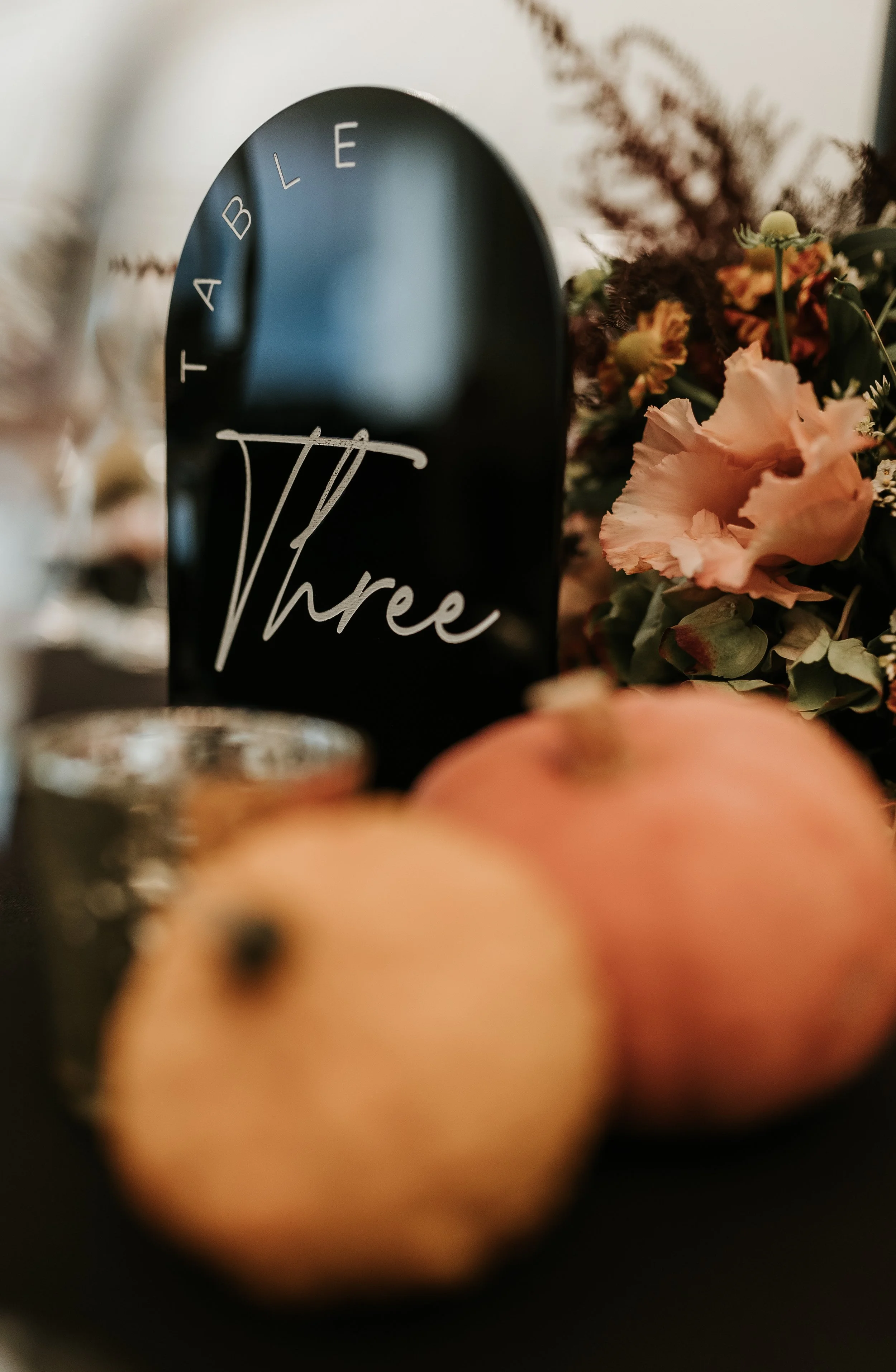 A black table number sign with the text "TABLE Three" beside pink flowers and peaches.