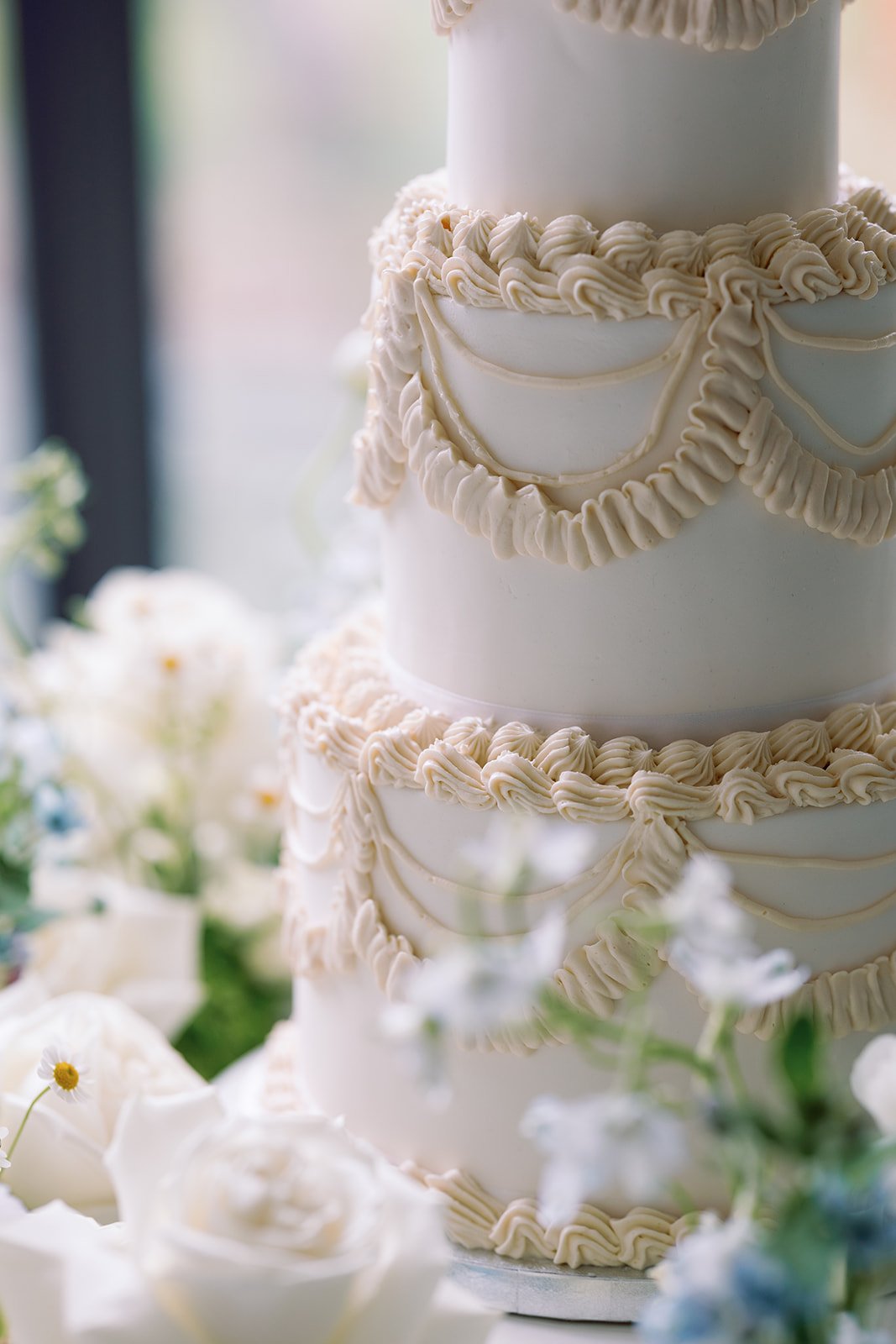 A multi-tier white wedding cake decorated with cream-colored piping and scrolls, surrounded by white flowers and greenery.