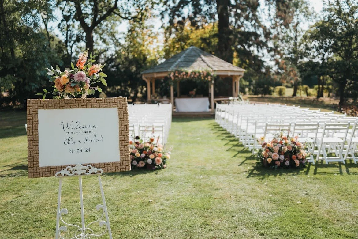 Outdoor wedding ceremony setup on a lawn with white chairs, floral arrangements, and a gazebo in the background, with a welcome sign for Ella and Michael's wedding.