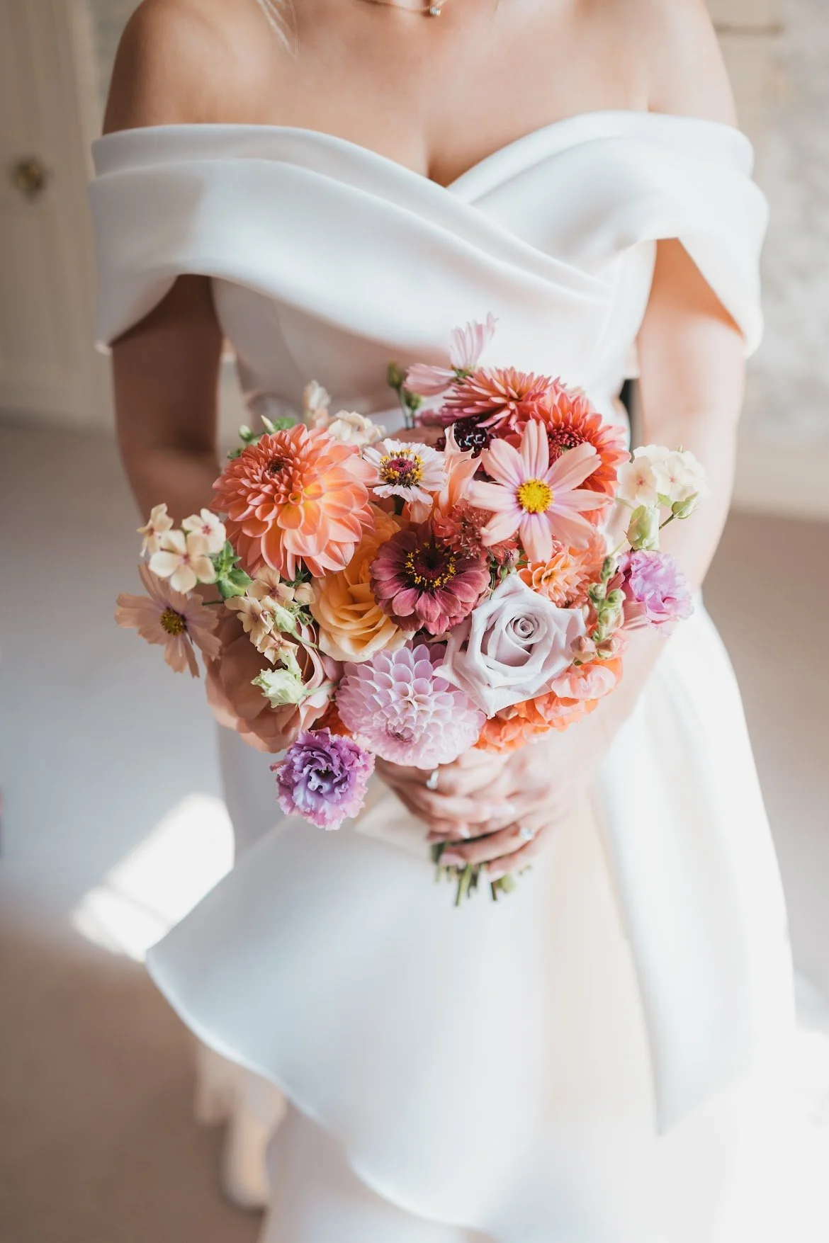 A woman in a white off-the-shoulder dress holding a colorful bouquet of various pink, peach, and purple flowers.