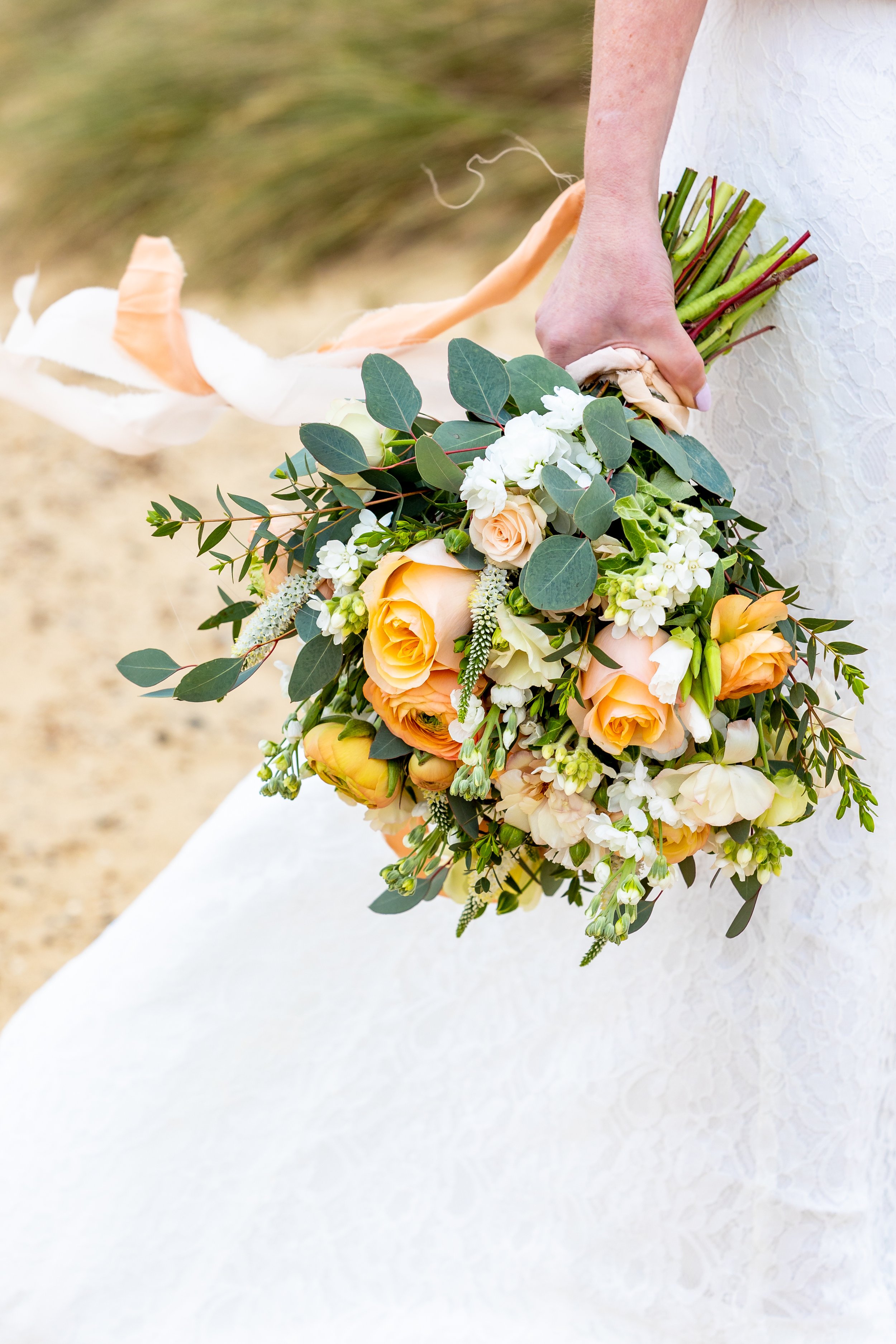 Bride holding a large bouquet of peach, white, and cream roses, surrounded by greenery, in a white wedding dress.