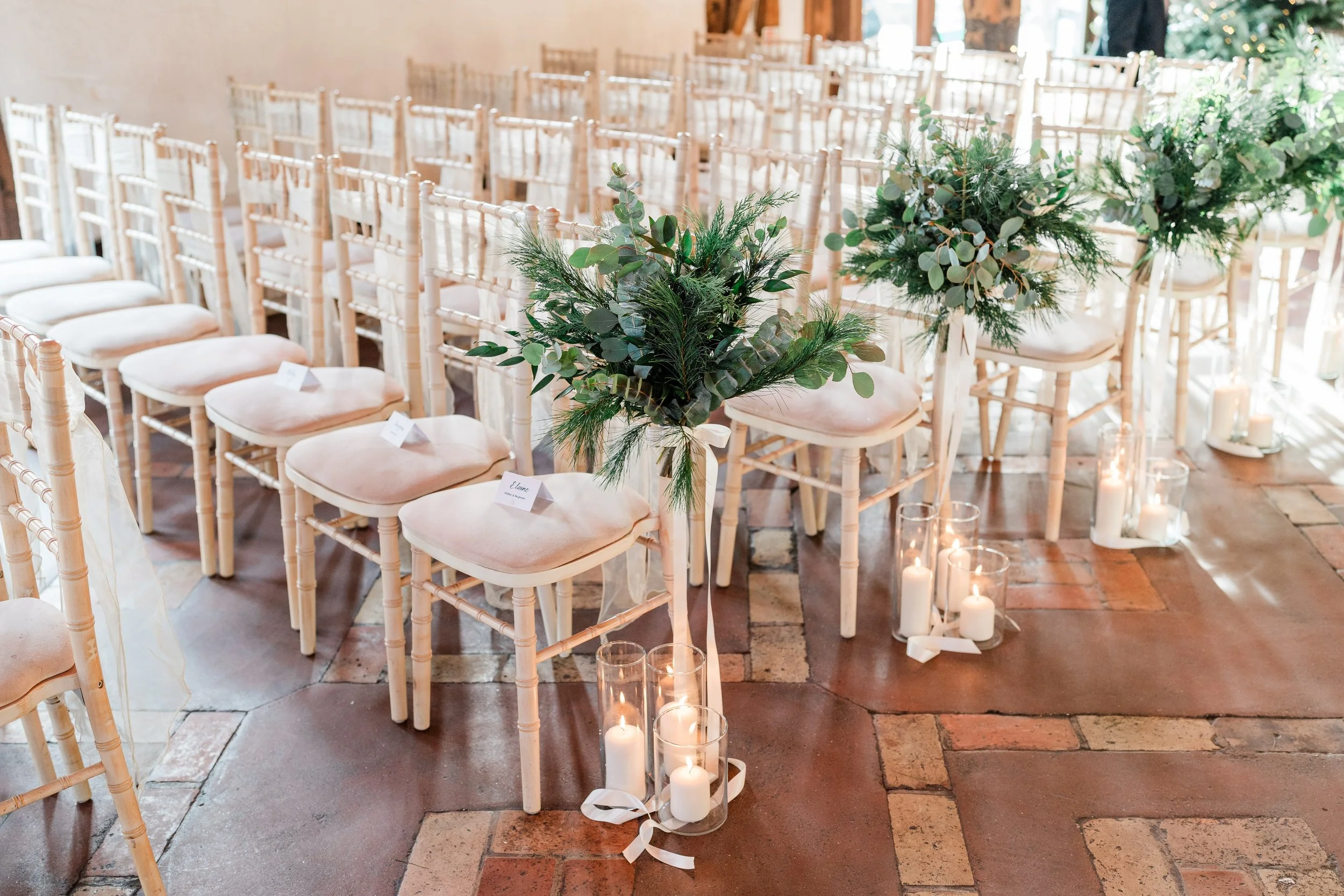 Elegant wedding ceremony setup with rows of white chairs, decorative green foliage on tall stands, and candles in glass holders on the floor.