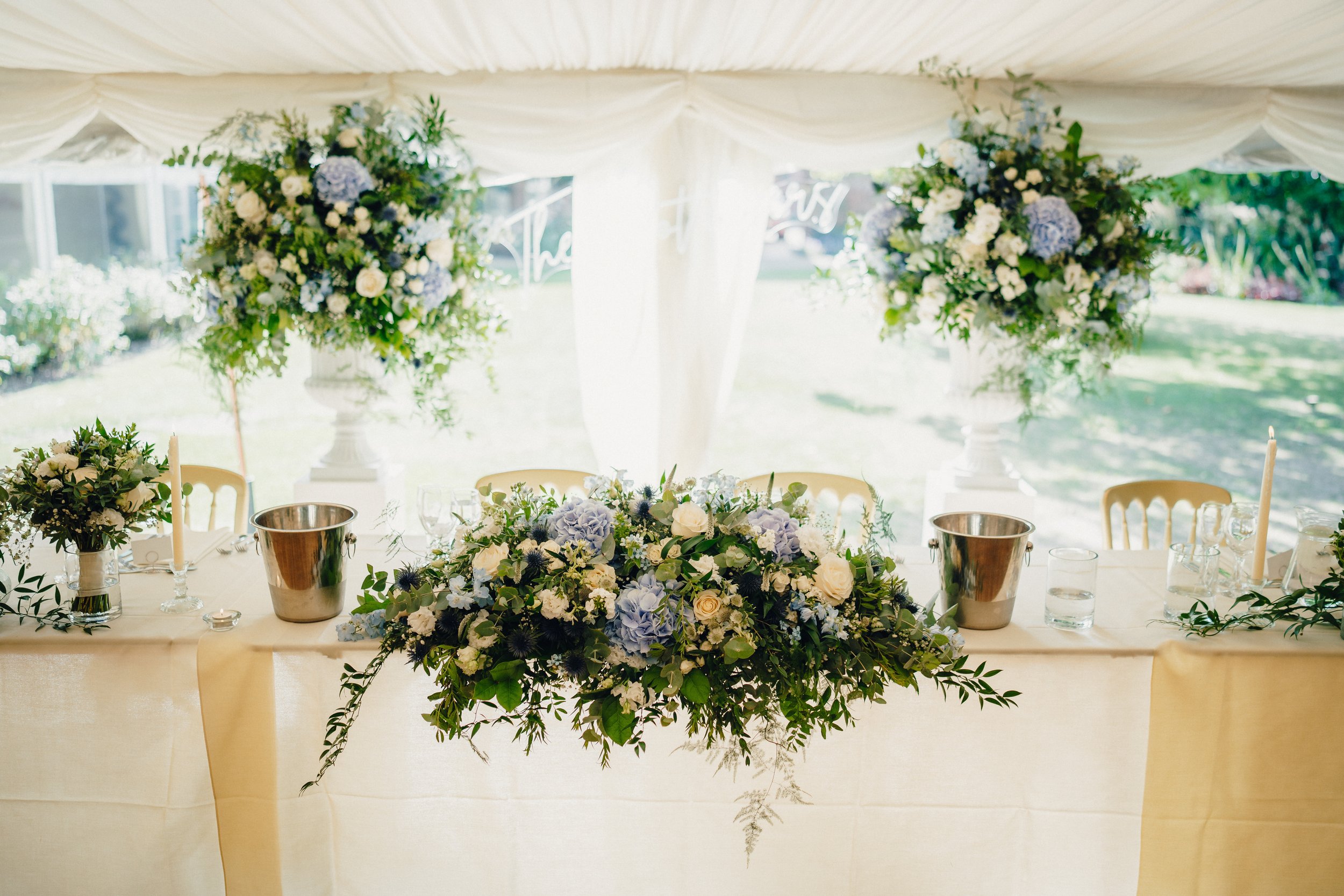Wedding reception table decorated with white, blue, and green floral arrangements, candles, and glassware inside a white tent with a garden outside.