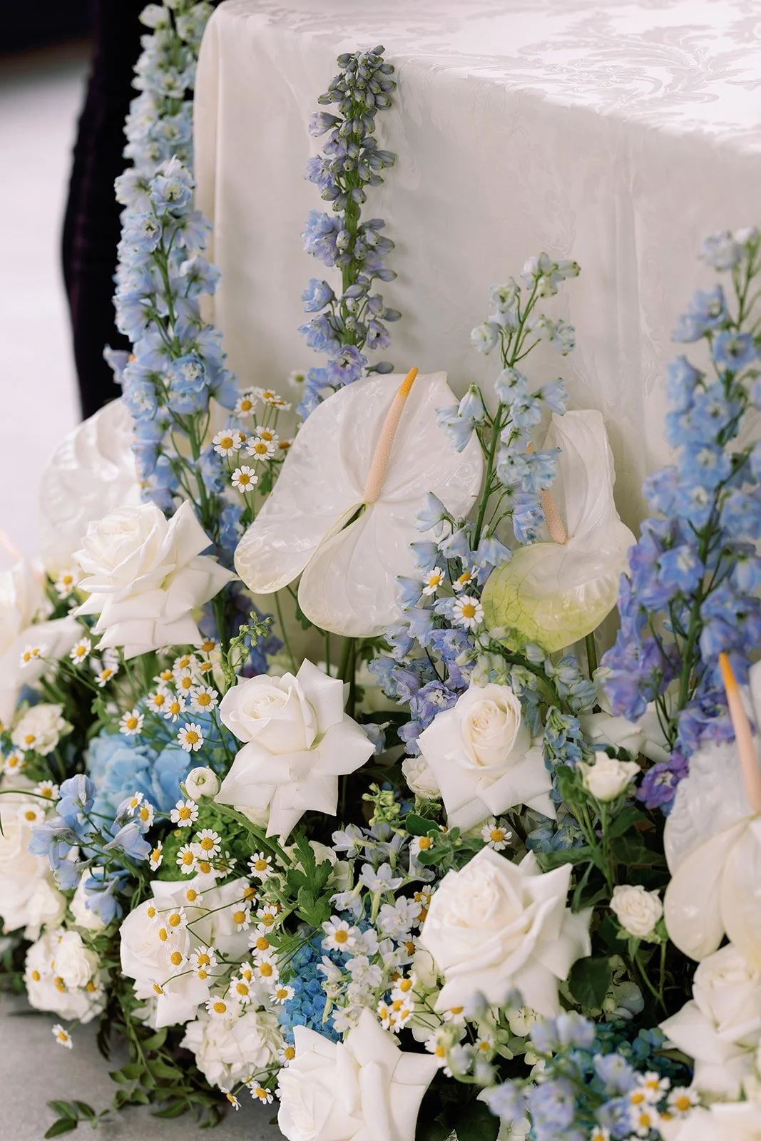 Flower arrangement with white roses, blue delphiniums, white calla lilies, and daisies, with an anthurium with a white spadix, at a table with white tablecloth.