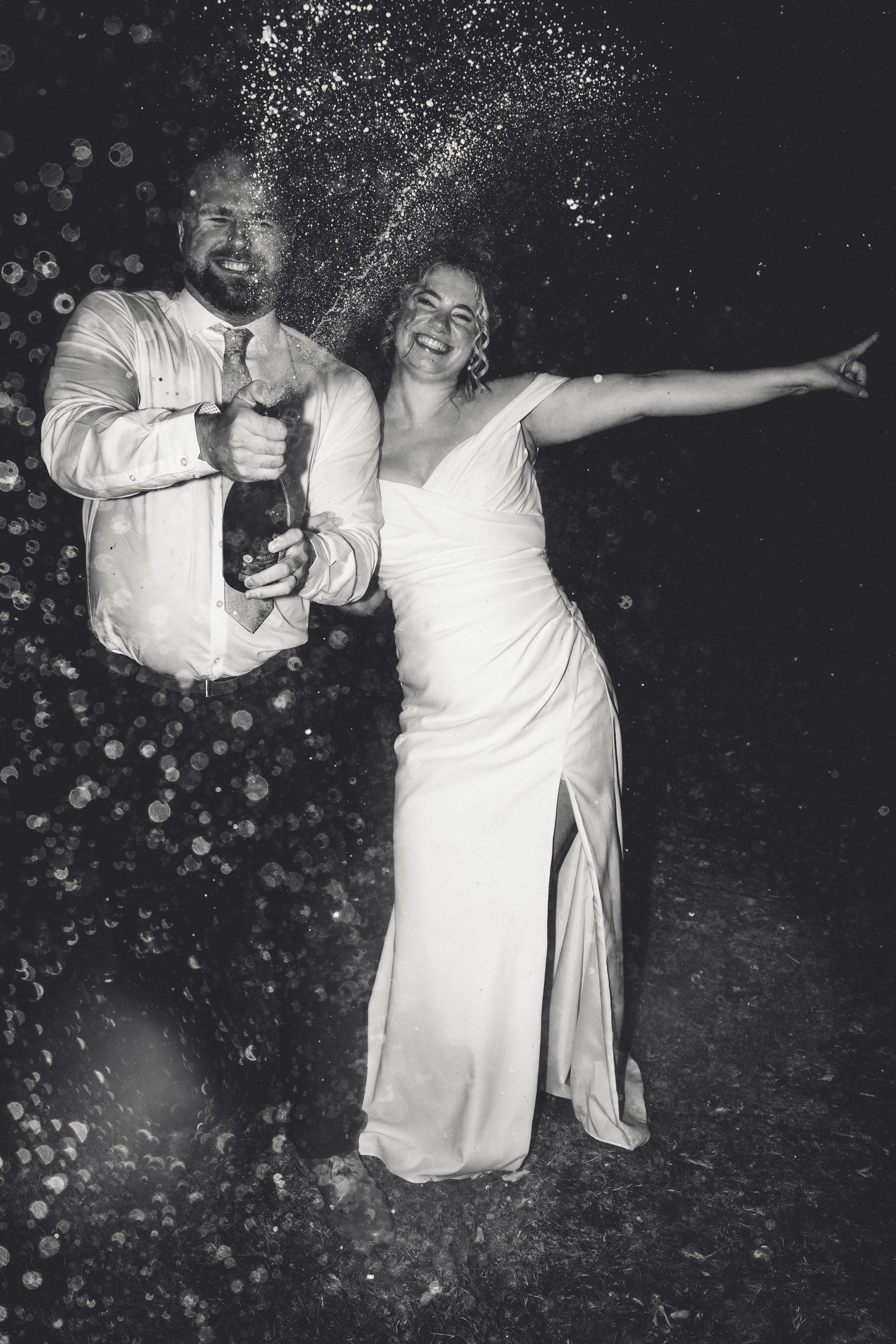 A black and white photo of a man and woman celebrating, with the man holding a champagne bottle and both smiling and laughing, fireworks or sparklers in the background.