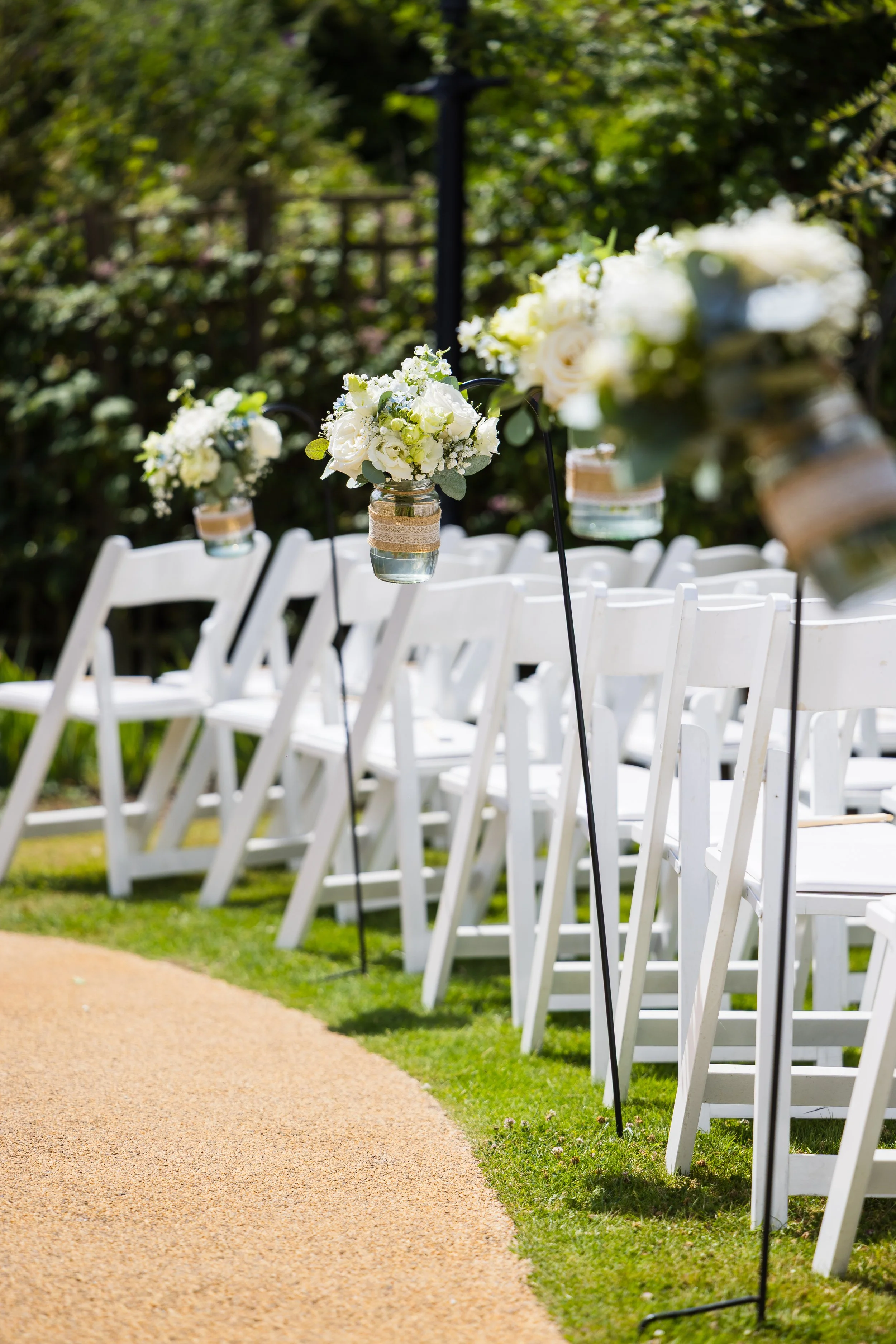 White chairs arranged outdoors along a pathway with hanging floral centerpieces featuring white roses and greenery.