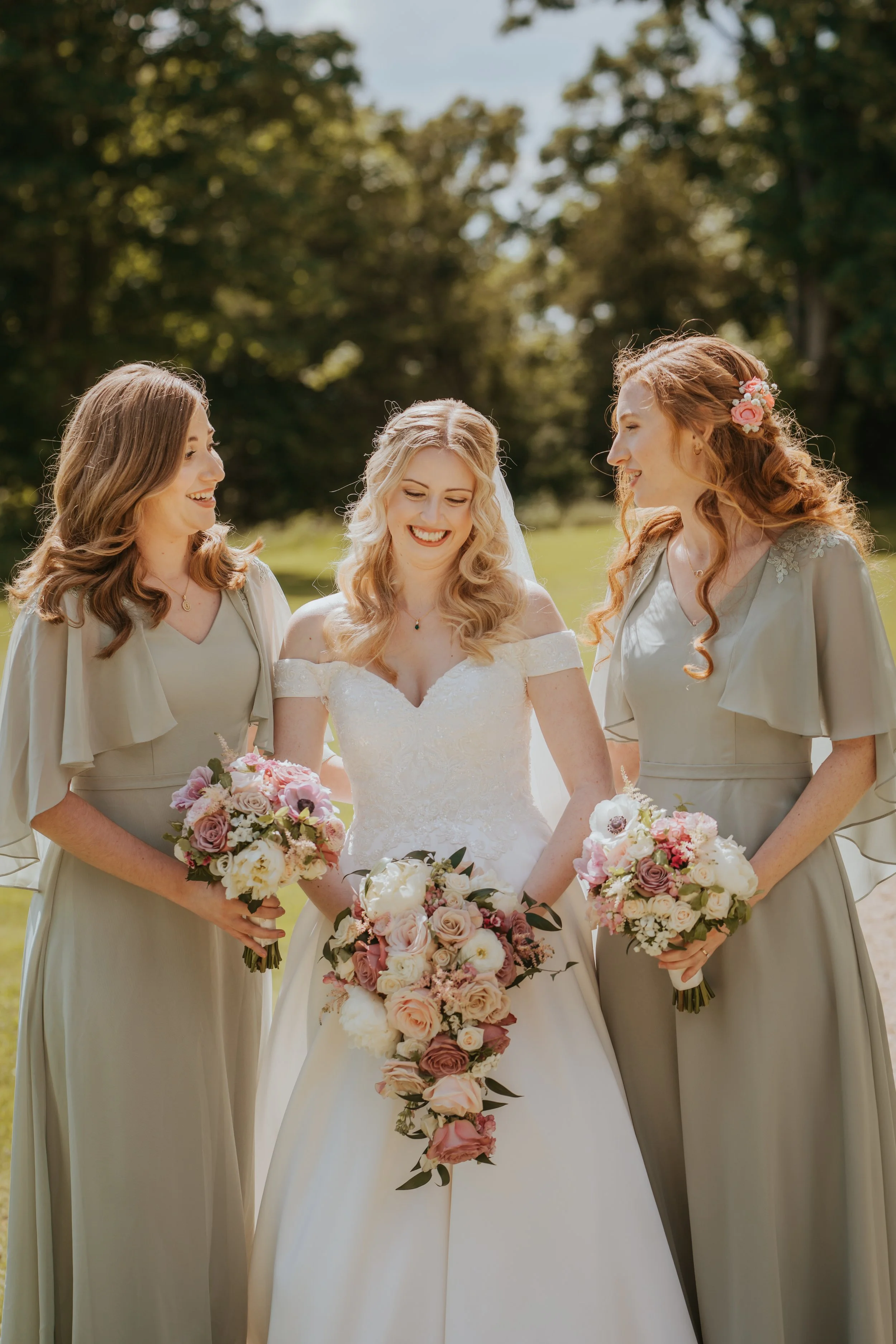 A bride with two bridesmaids outdoors, all holding bouquets and smiling.