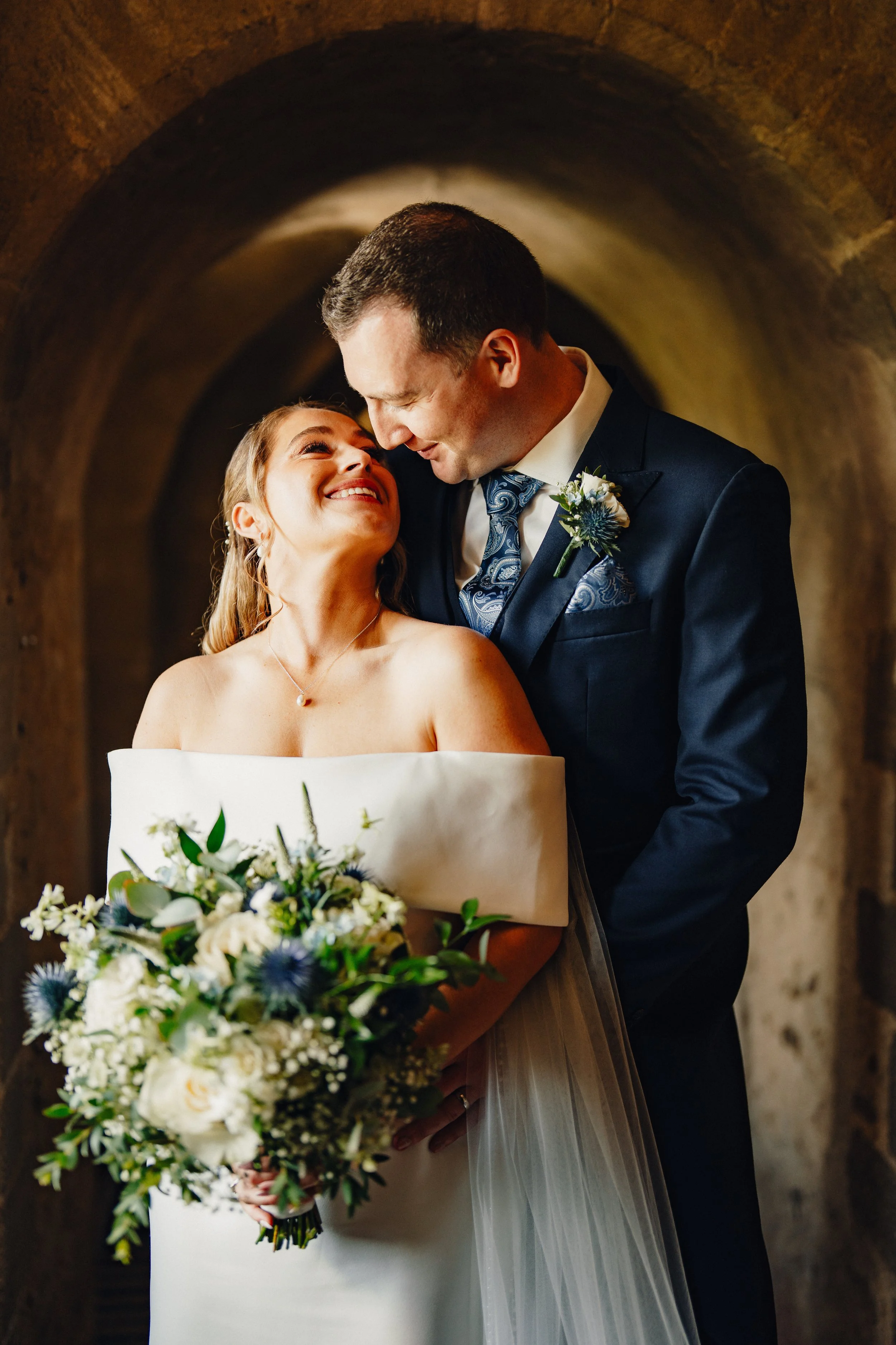 A bride and groom share a loving moment during their wedding, standing close together in a brick archway, with the bride holding a bouquet of white and blue flowers.