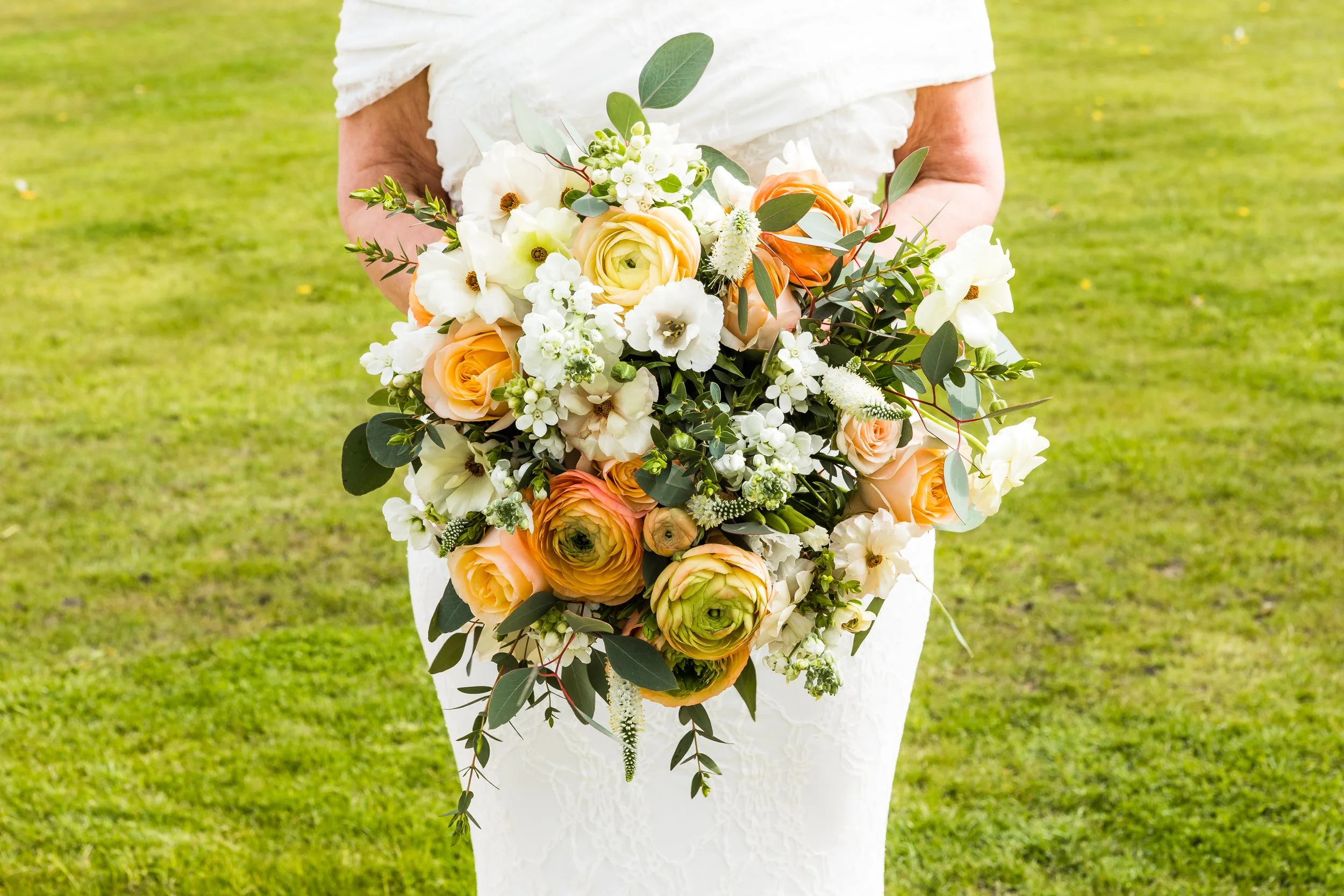 A person in a white dress holding a large bouquet of white, peach, and orange flowers with green leaves outdoors on grass.