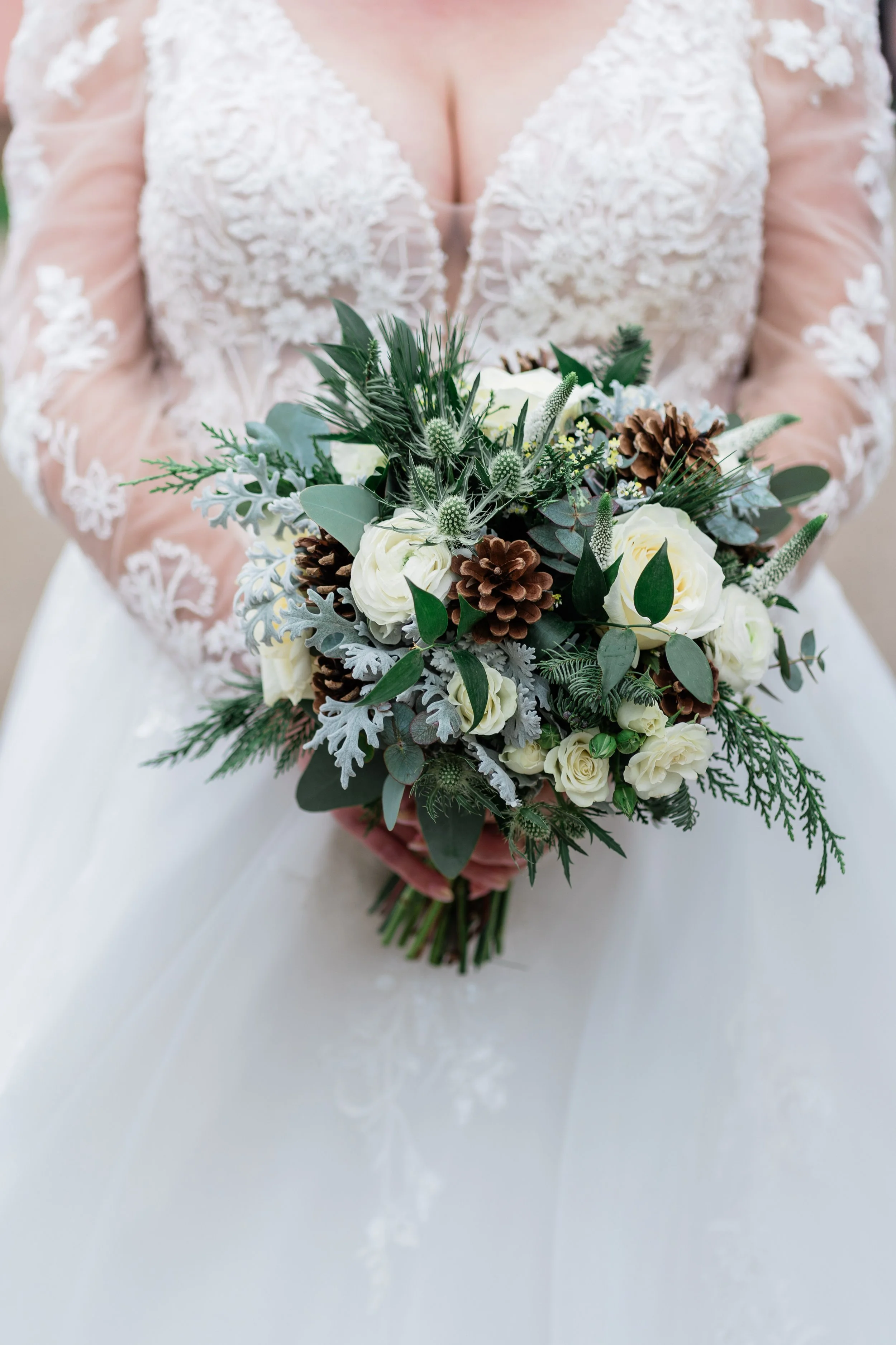 A bride in a lace wedding dress holding a bouquet of white roses, pinecones, and greenery.