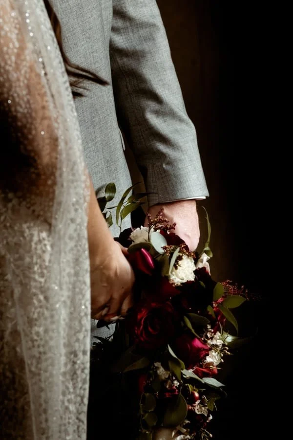 A bride and groom holding a bouquet, with the bride in a lace dress and the groom in a gray suit, against a dark background.